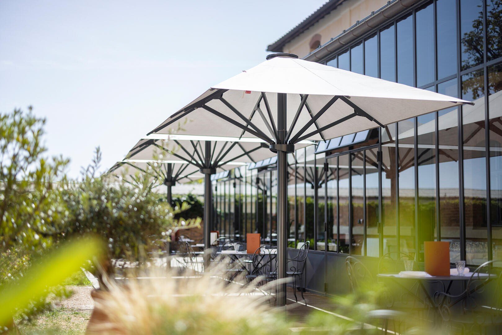 Sitzgelegenheiten im Außenbereich eines Restaurants mit Metalltischen und -stühlen unter großen weißen Sonnenschirmen neben einem modernen Glasgebäude, umgeben von Grün und Sonnenlicht.