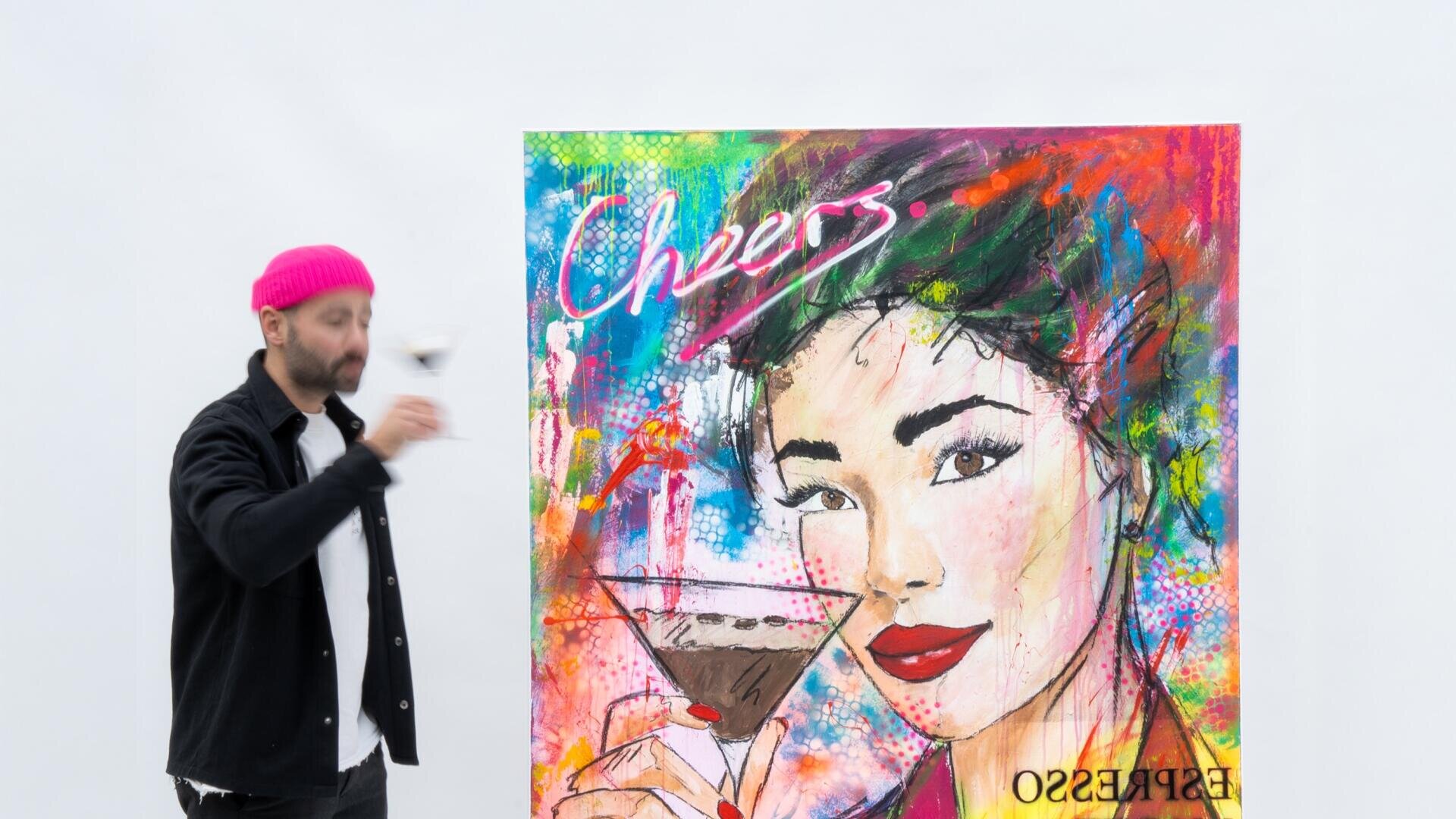Ein Mann mit einer rosa Mütze hält ein Martini-Glas in der Hand, neben einem farbenfrohen Gemälde, das eine Frau mit einem Martini zeigt; auf dem Bild sind die Worte „Cheers“ und „Espresso Martini“ zu lesen. Daneben stehen eine Flasche und ein Shaker.