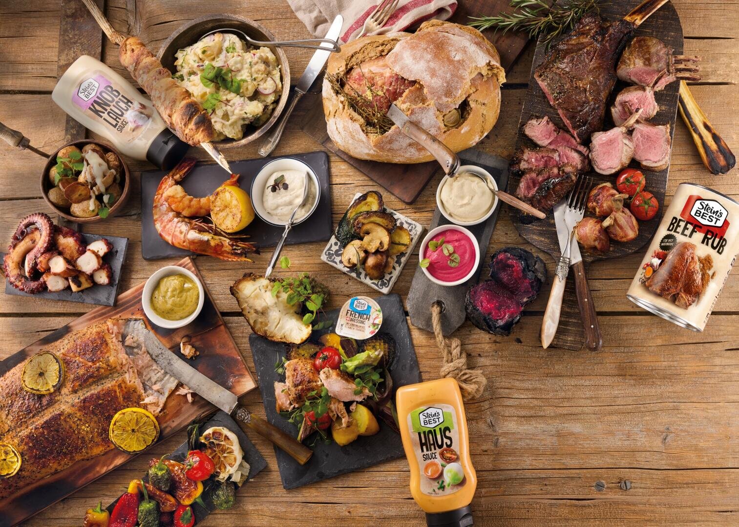 Eine Studioaufnahme aus hohem Blickwinkel eines rustikalen Holztisches, der mit einem Gourmet-Buffet gedeckt ist, darunter gegrillte Steaks, Lachs, Garnelen, Tintenfisch, gebratenes Gemüse, Kartoffelsalat, handwerklich hergestelltes Brot sowie verschiedene Steins Best-Saucen und Trockenmarinaden.