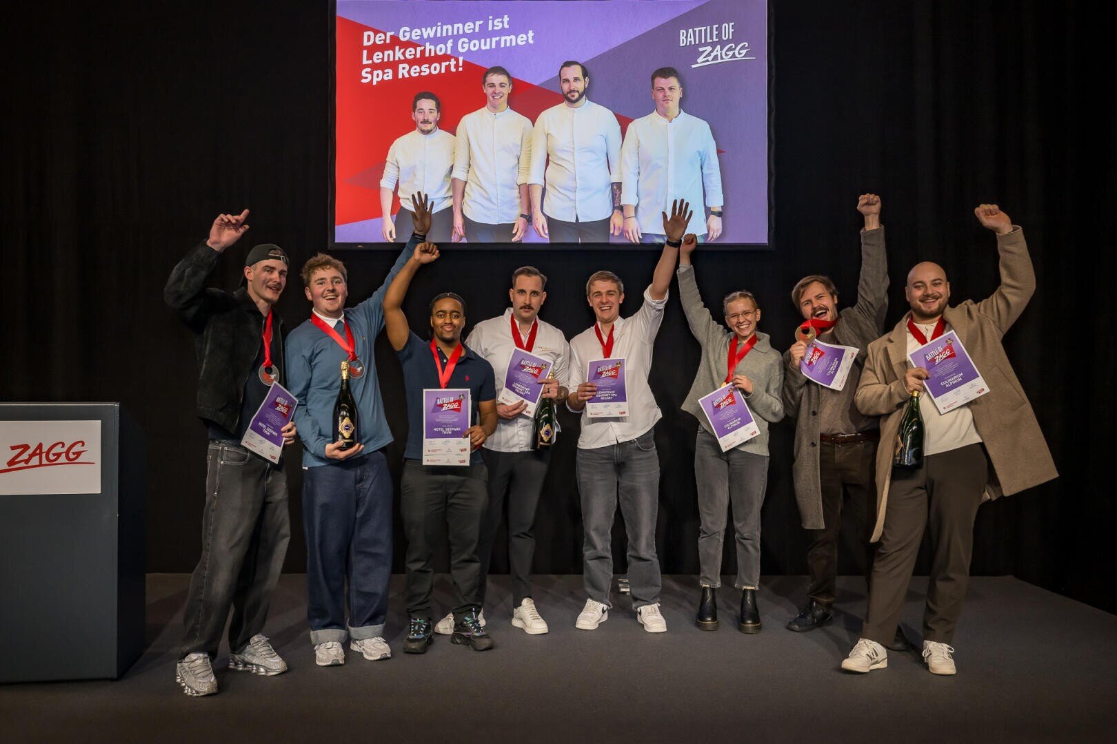 Acht Personen feiern auf einer Bühne mit Auszeichnungen und Champagner in der Hand vor einer Leinwand, auf der das Lenkerhof Gourmet Spa Resort als Gewinner der Battle of ZAGG verkündet wird.