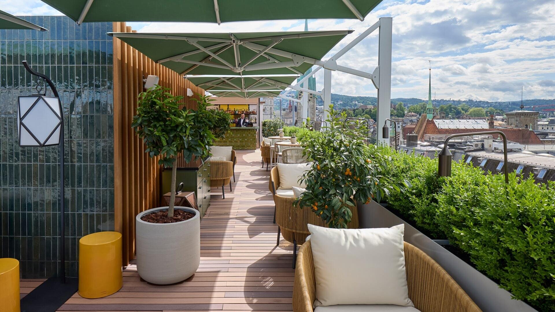 Dachterrasse mit gepolsterten Stühlen, Topfpflanzen, grünen Sonnenschirmen und Blick auf die Stadt an einem sonnigen Tag. Holzboden und grüne Hecken tragen zur gemütlichen, modernen Atmosphäre bei.