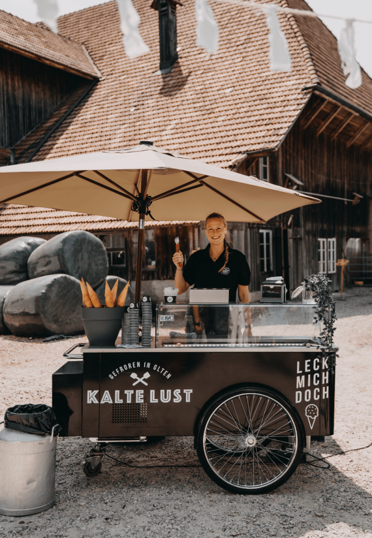 Eine Person steht lächelnd hinter einem Eiswagen mit einem Sonnenschirm vor rustikalen Gebäuden. Auf dem Wagen steht „Kalte Lust“, daneben liegen Baguettes, Eistüten und ein Mülleimer.