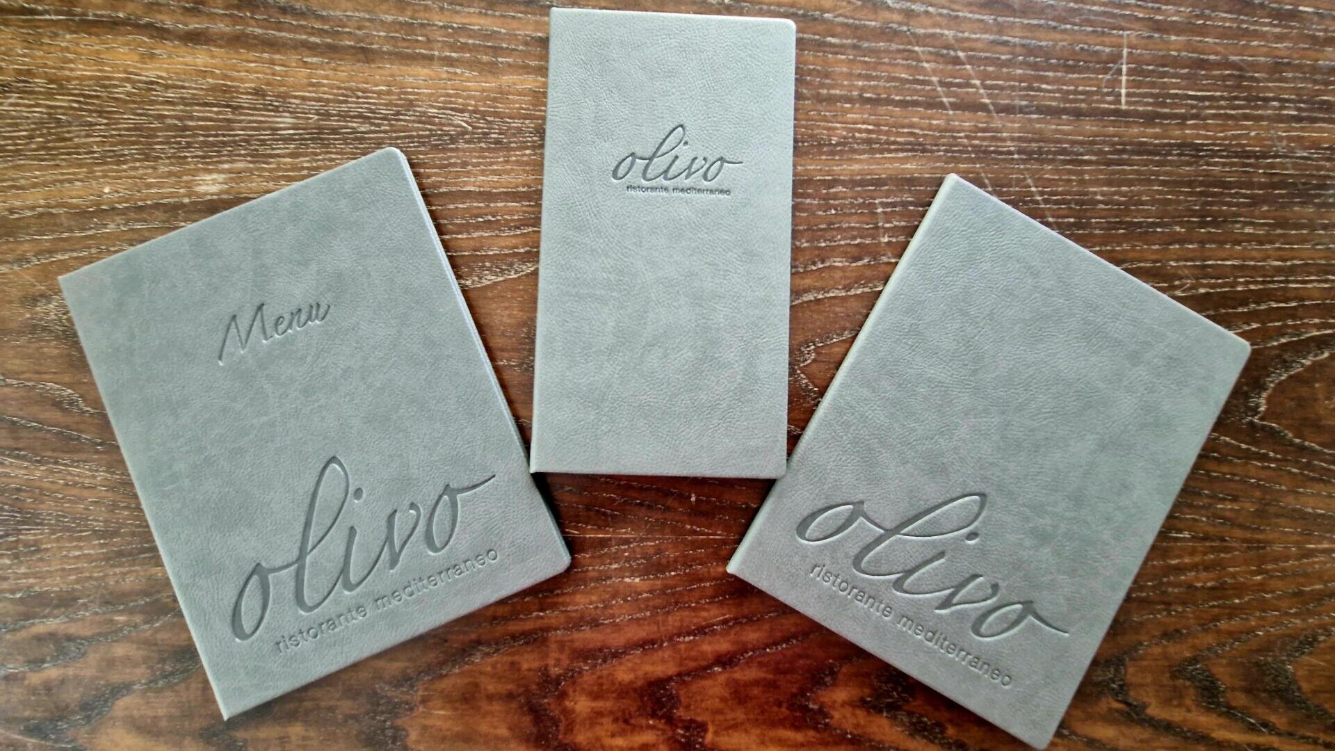 Auf einem Holztisch liegen drei graue Speisekartenhefte, auf deren Einbänden „olivo ristorante mediterraneo“ eingeprägt ist; zwei davon sind aufgeschlagen, eines liegt geschlossen in der Mitte.