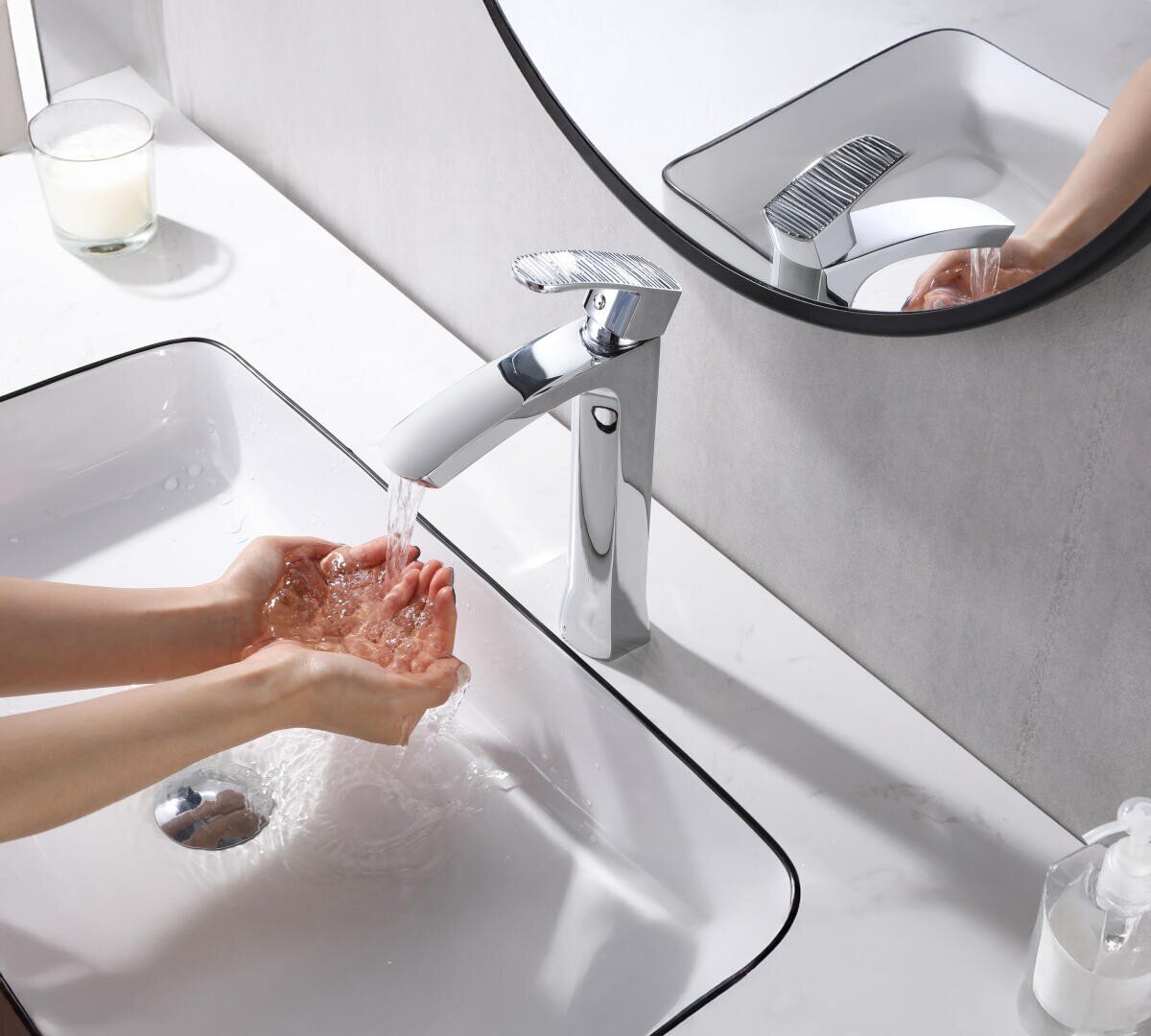 Eine Person wäscht sich die Hände unter einem modernen verchromten Wasserhahn in einem weißen Waschbecken, wobei das Wasser spritzt. Ein runder Spiegel, ein Seifenspender und eine Glaskerze stehen auf der weißen Arbeitsplatte.