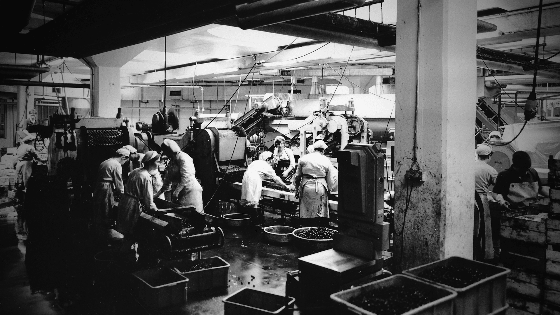 Schwarz-Weiß-Foto einer belebten Fabrikhalle, in der Arbeiter in Uniformen und mit Hüten Waren an großen Industriemaschinen verarbeiten, mit Behältern, Rohren und Kisten, die überall im Arbeitsbereich verstreut sind.