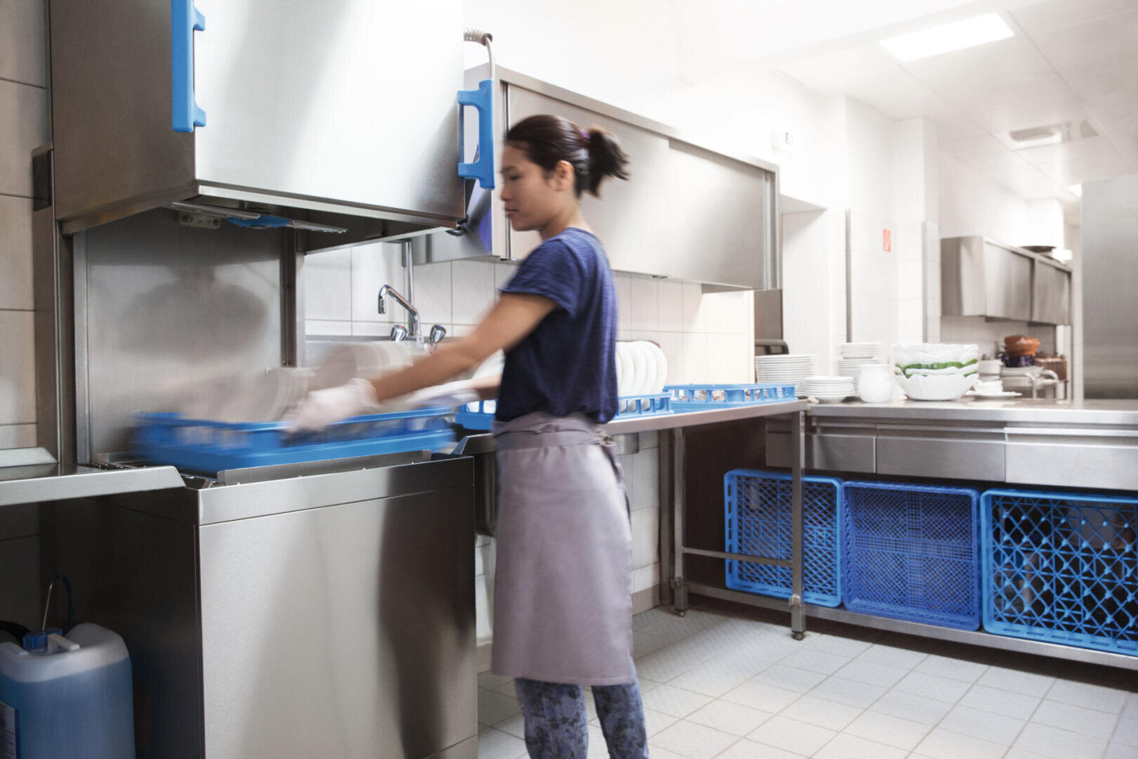 Eine Person, die eine Schürze trägt, stellt einen blauen Geschirrkorb in eine gewerbliche Haubenspülmaschine der Marke MEIKO M-iClean in einer hellen, sauberen Küche. Im Hintergrund sind Stapel von Tellern und weitere blaue Körbe zu sehen.