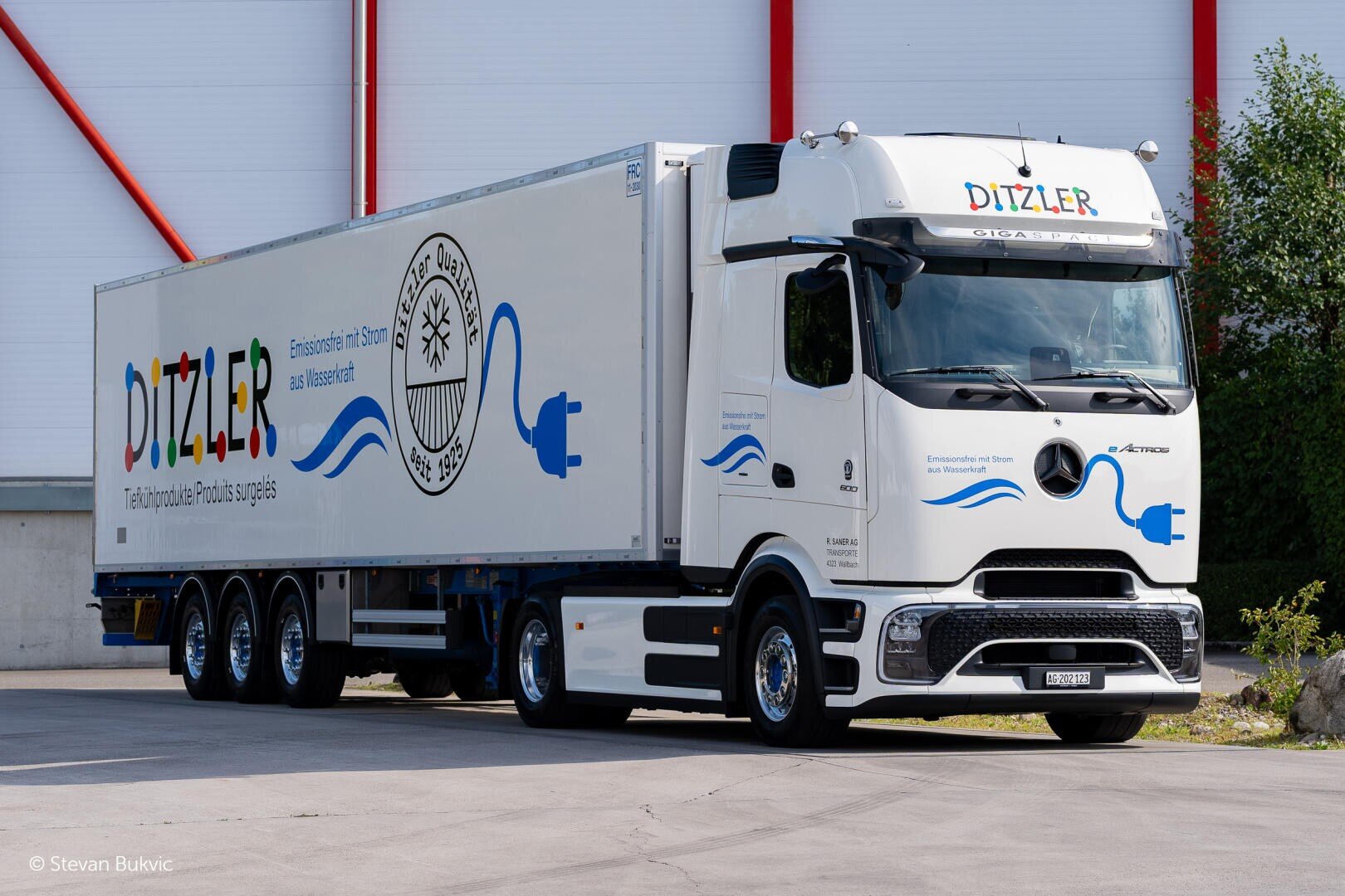 Ein weißer Elektro-Sattelzug mit DITZLER-Schriftzug und Elektrostecker-Grafik ist vor einem Gebäude geparkt, mit einem Mercedes-Benz-Logo auf der Vorderseite und einem großen Anhänger daran.