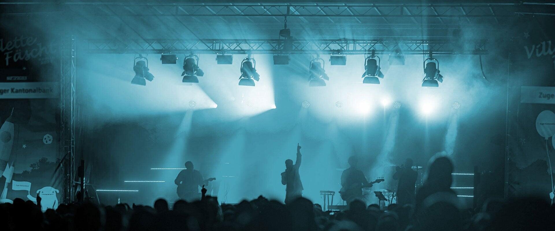 Eine Band tritt auf einer verrauchten, blau beleuchteten Bühne vor einem großen Publikum auf. Der Leadsänger steht in der Mitte und hebt einen Arm, während die anderen Musiker im Hintergrund ihre Instrumente spielen.