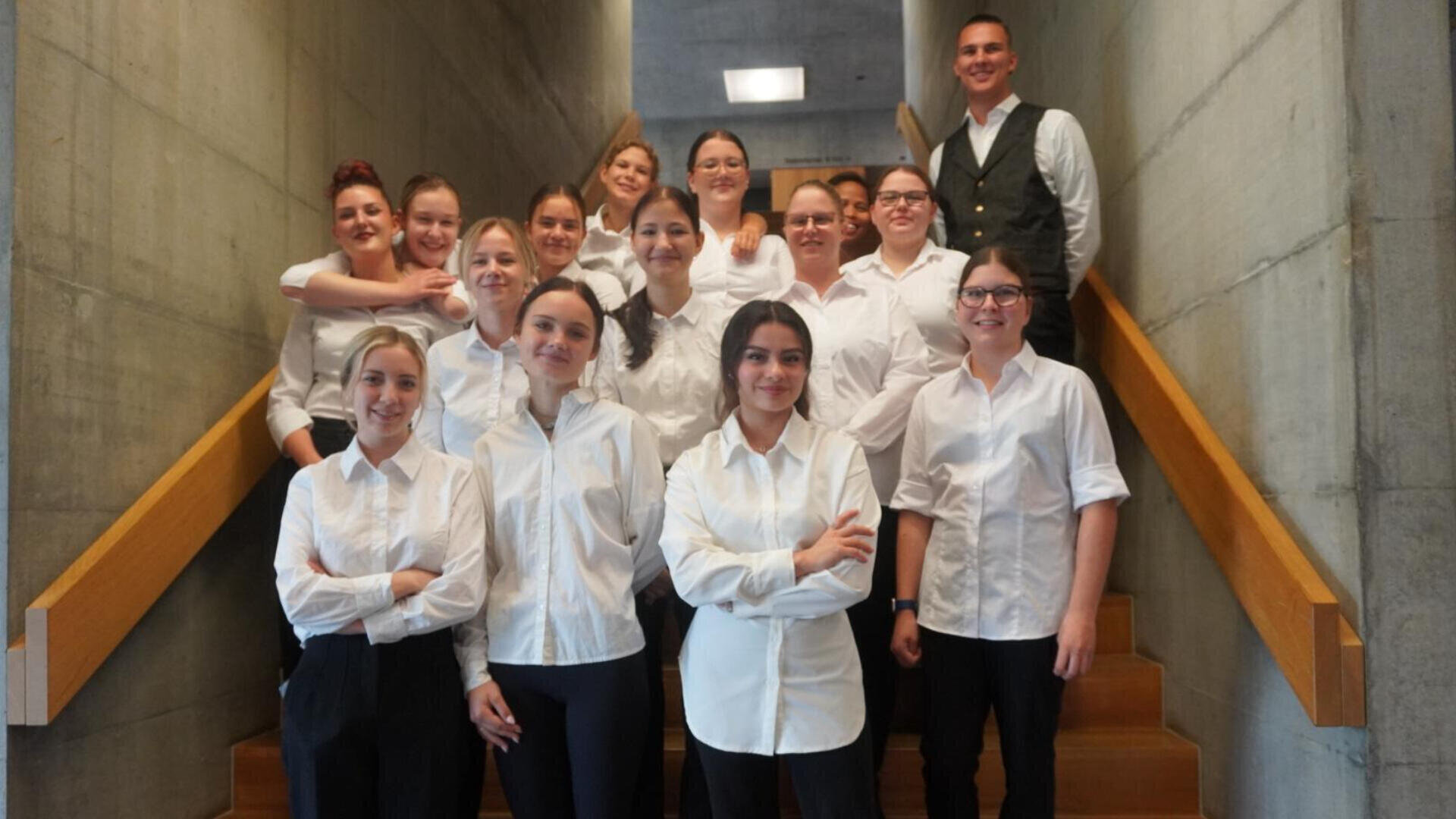 Eine Gruppe von dreizehn Personen in weißen Hemden steht lächelnd auf einer Treppe zwischen Betonwänden; eine Person im Hintergrund trägt eine Weste über dem Hemd.