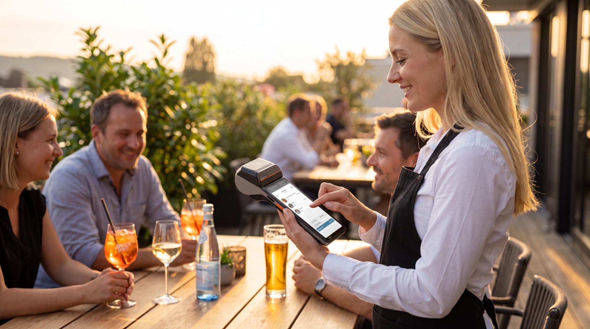 Ein lächelnder Kellner bedient an einem Tisch im Außenbereich eines Restaurants, an dem drei Personen an einem sonnigen Tag bei einem Getränk die Unterhaltung genießen, mit einem mobilen Zahlungsgerät.