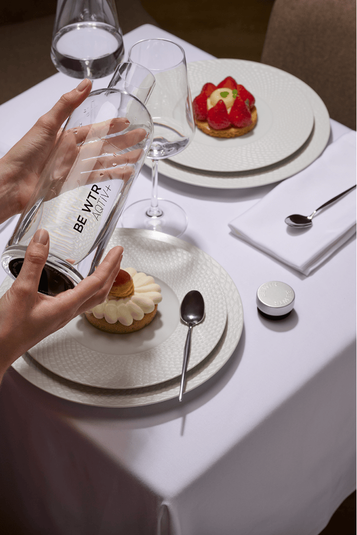 Eine Person gießt Wasser aus einer Glasflasche mit der Aufschrift „BE MY TREAT AQUA+“ auf einen Tisch, der mit weißen Tellern, zwei Stück Gebäck, Gläsern, Löffeln und einer weißen Serviette gedeckt ist.