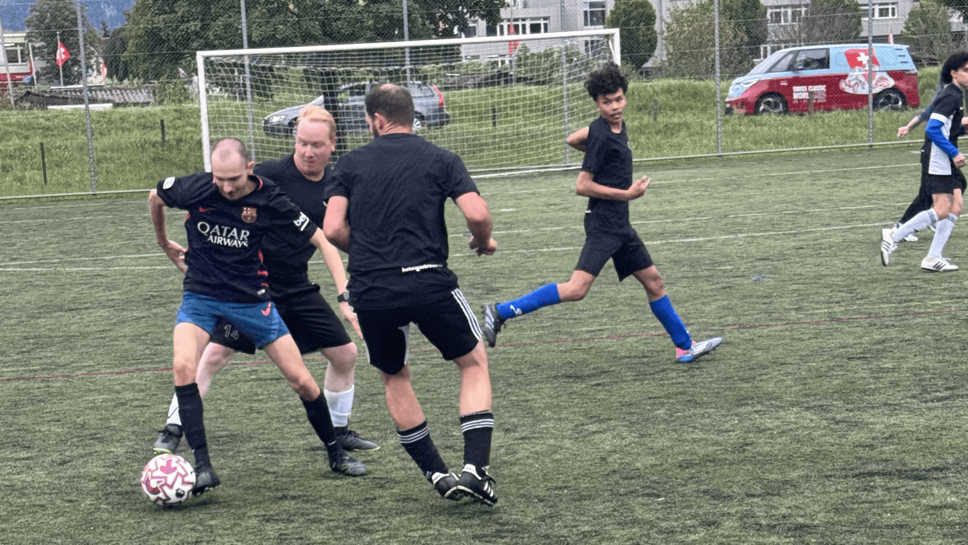 Vier Männer spielen Fußball auf einem grünen Feld; zwei kämpfen um den Ball, während die anderen in der Nähe herumlaufen. Im Hintergrund sind ein weißes Tor und ein rotes Auto zu sehen. Alle Spieler tragen schwarze Trikots sowie unterschiedliche Shorts und Socken.