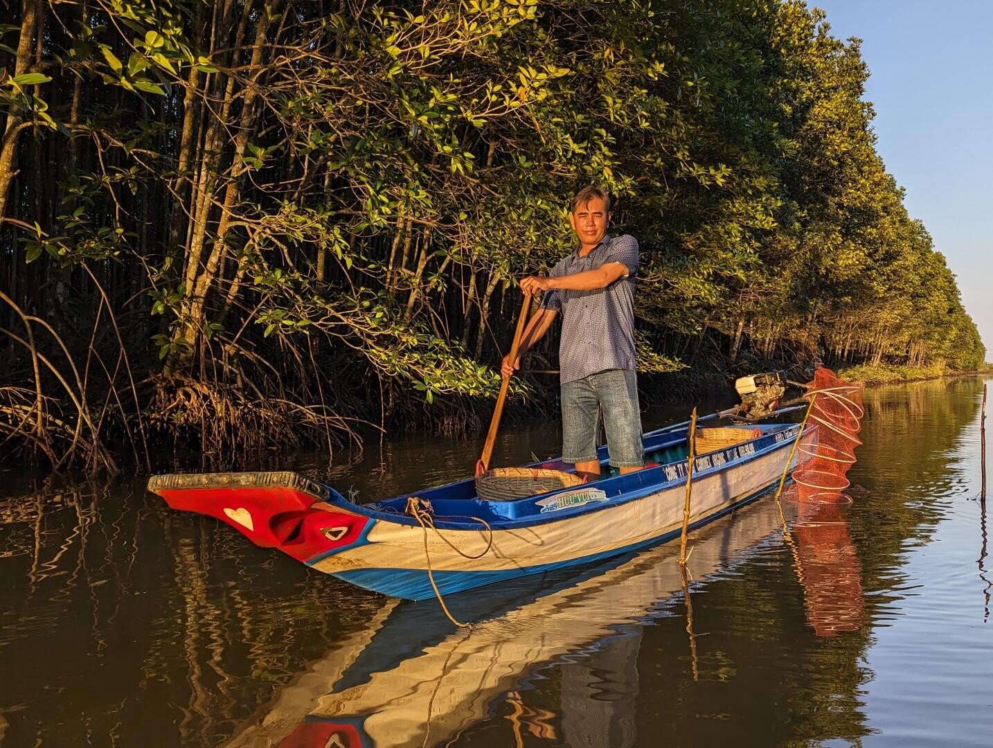Eine Person steht in einem blauen Holzboot auf einem ruhigen Fluss und paddelt, umgeben von dichten grünen Bäumen und sonnenbeschienenem Laub, das sich im Wasser spiegelt.
