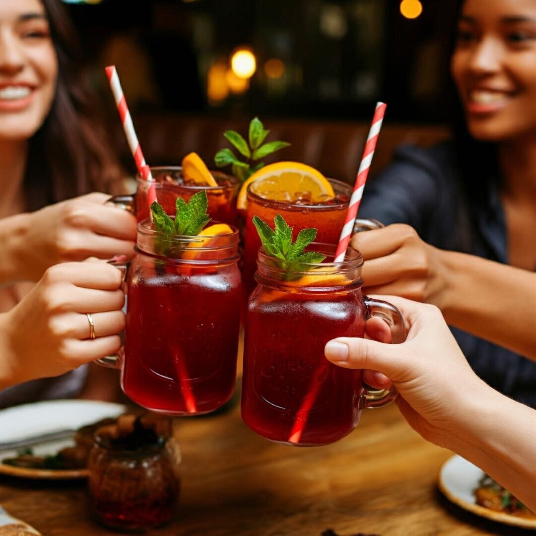 Drei Personen stoßen mit gestreiften Strohhalmen an Gläsern an, die mit einem roten Getränk gefüllt sind, das mit Orangenscheiben und Minze garniert ist. Sie lächeln und sitzen an einem Holztisch in einem gemütlichen Restaurant.