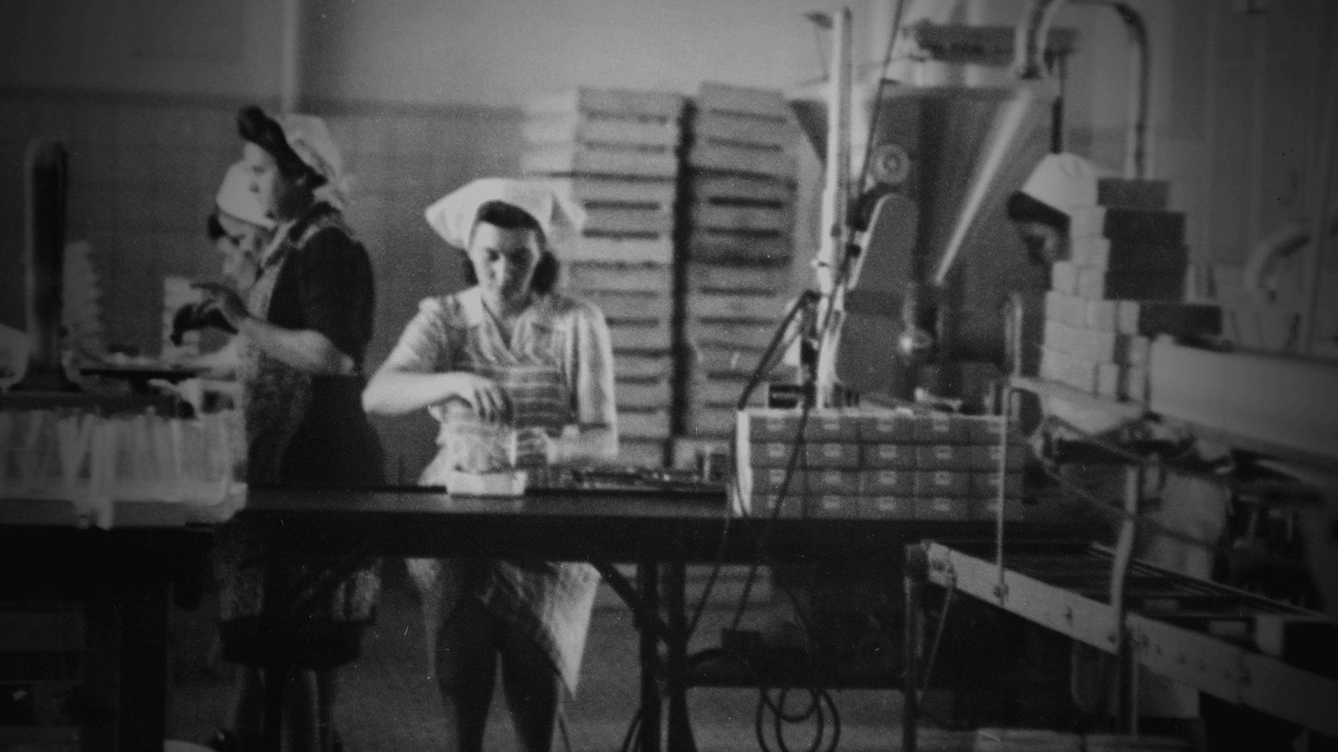 Schwarz-Weiß-Foto von Frauen, die in einer Fabrik arbeiten, Artikel an Tischen verpacken und Kopftücher oder Haarbedeckungen tragen, mit Stapeln von Kisten und Industrieanlagen im Hintergrund.
