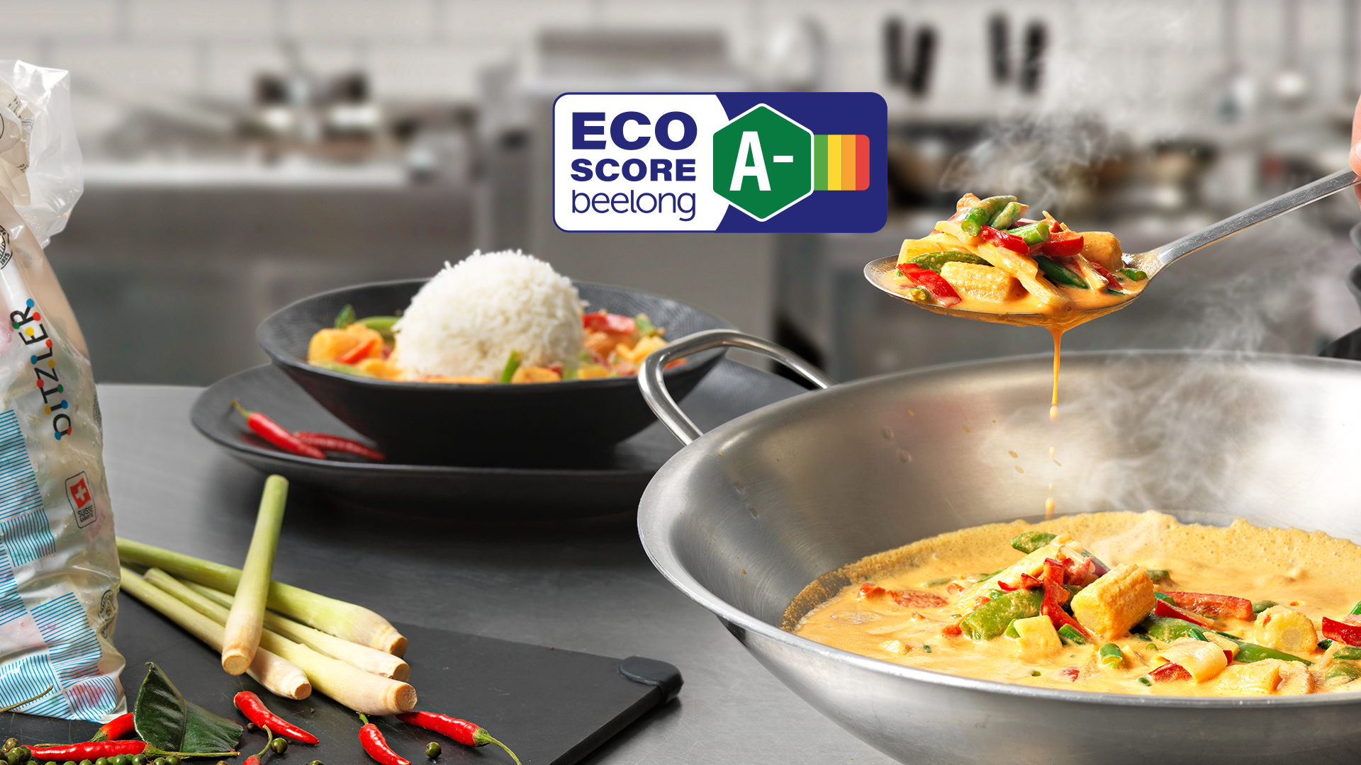 Eine Pfanne gelbes Curry mit Thai-Mix Gemüsemischung wird mit Reis serviert. Frische Zutaten wie Zitronengras und Chili liegen daneben. Ein Eco-Score Label mit der Bewertung A- ist zu sehen.