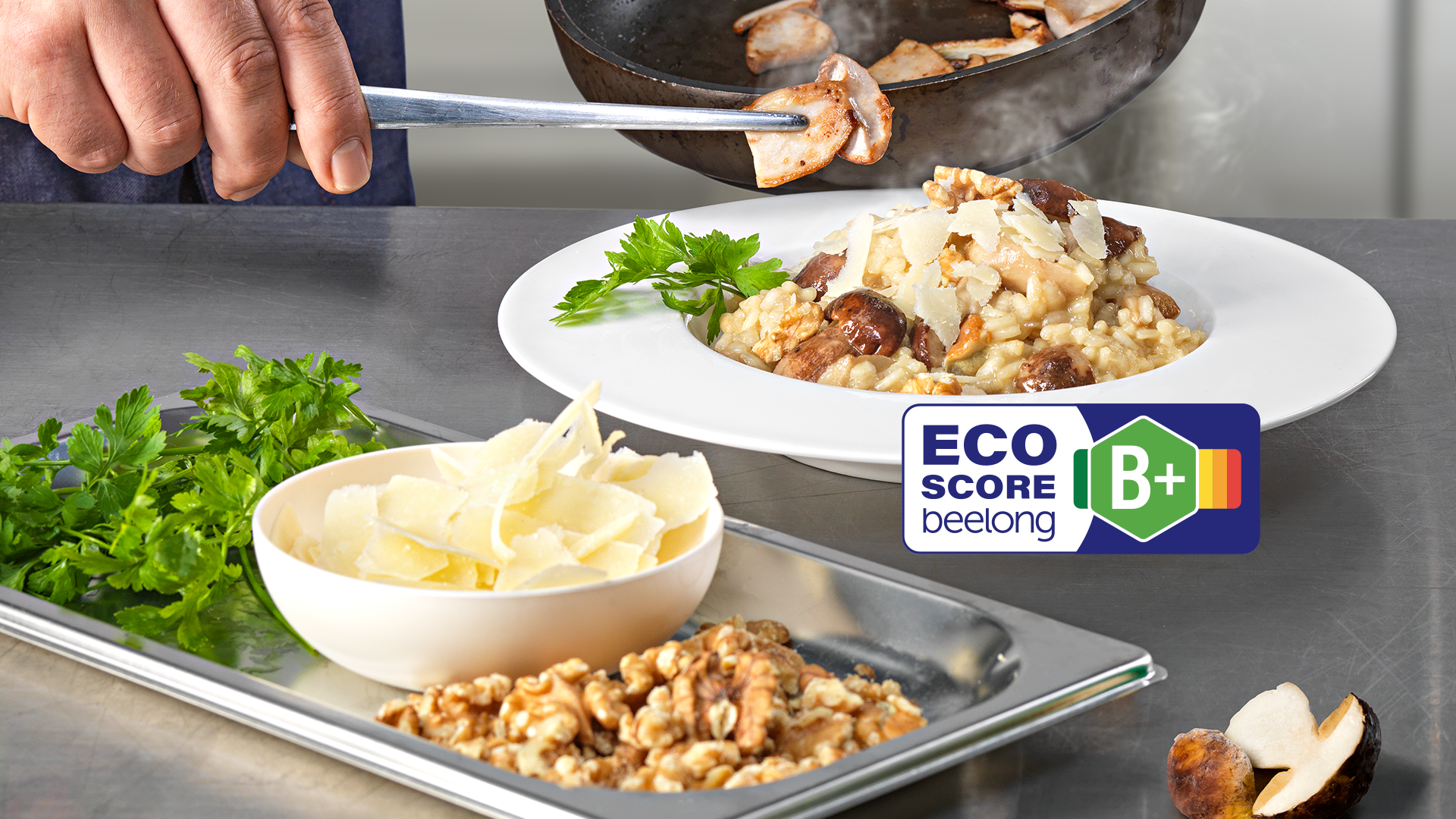 Eine Hand gibt gekochte Steinpilze aus der Pfanne auf einen Teller Risotto mit Käse und Petersilie. Frische Kräuter, Walnüsse und Käse liegen daneben. Im Vordergrund ist ein Eco-Score Label mit der Bewertung B+ zu sehen.