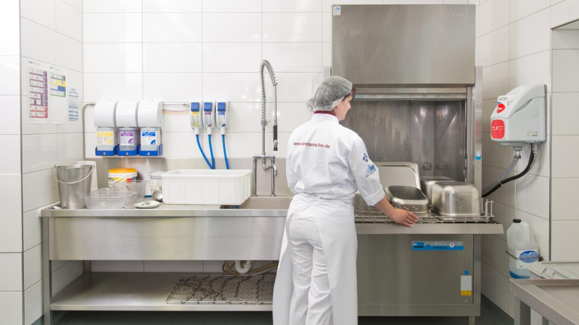 Eine Person in weißer Arbeitskleidung und mit Haarnetz lädt Metalltabletts in eine gewerbliche Spülmaschine der Marke MEIKO, Model FV 130.2, in einer sauberen, gefliesten Küche. An der linken Wand sind Seifenspender und ein Spülbecken zu sehen.