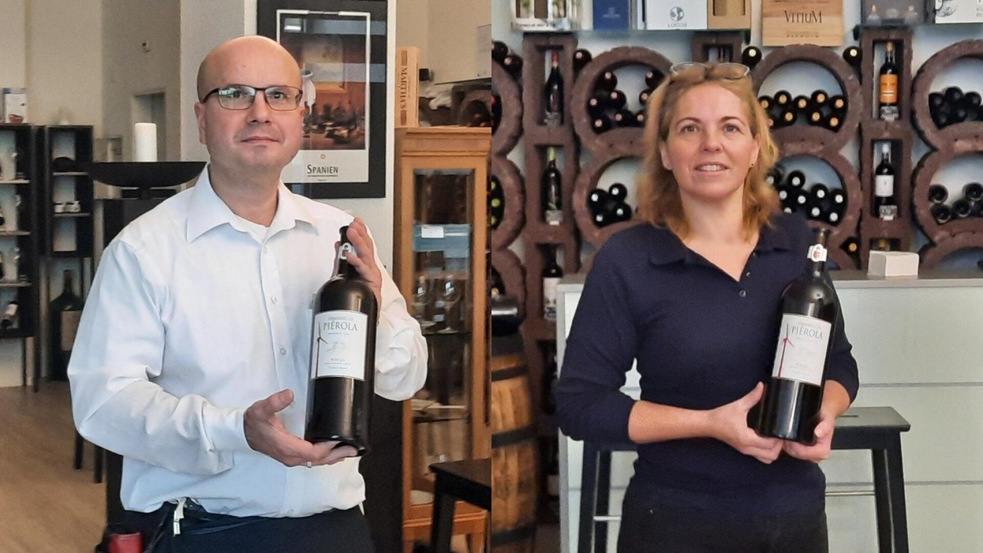 Ein Mann und eine Frau stehen in einem Weinladen und halten jeweils eine große Weinflasche in der Hand. Im Hintergrund sind Regale mit Weinflaschen und Holzdekorationen zu sehen.