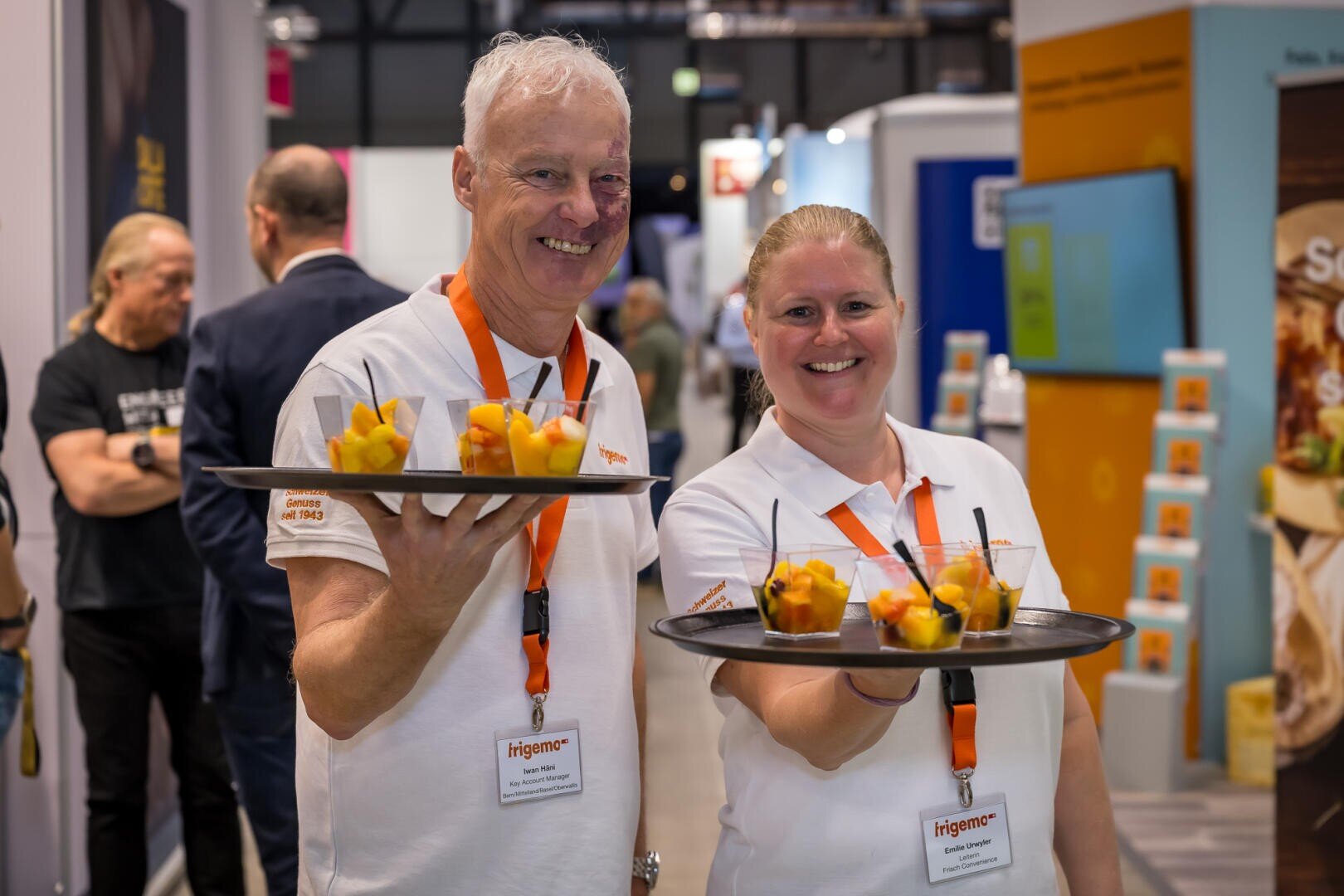 Ein Mann und eine Frau in weißen Polohemden mit Logos, die auf einer Messe lächelnd Tabletts mit Obstbechern in den Händen halten.