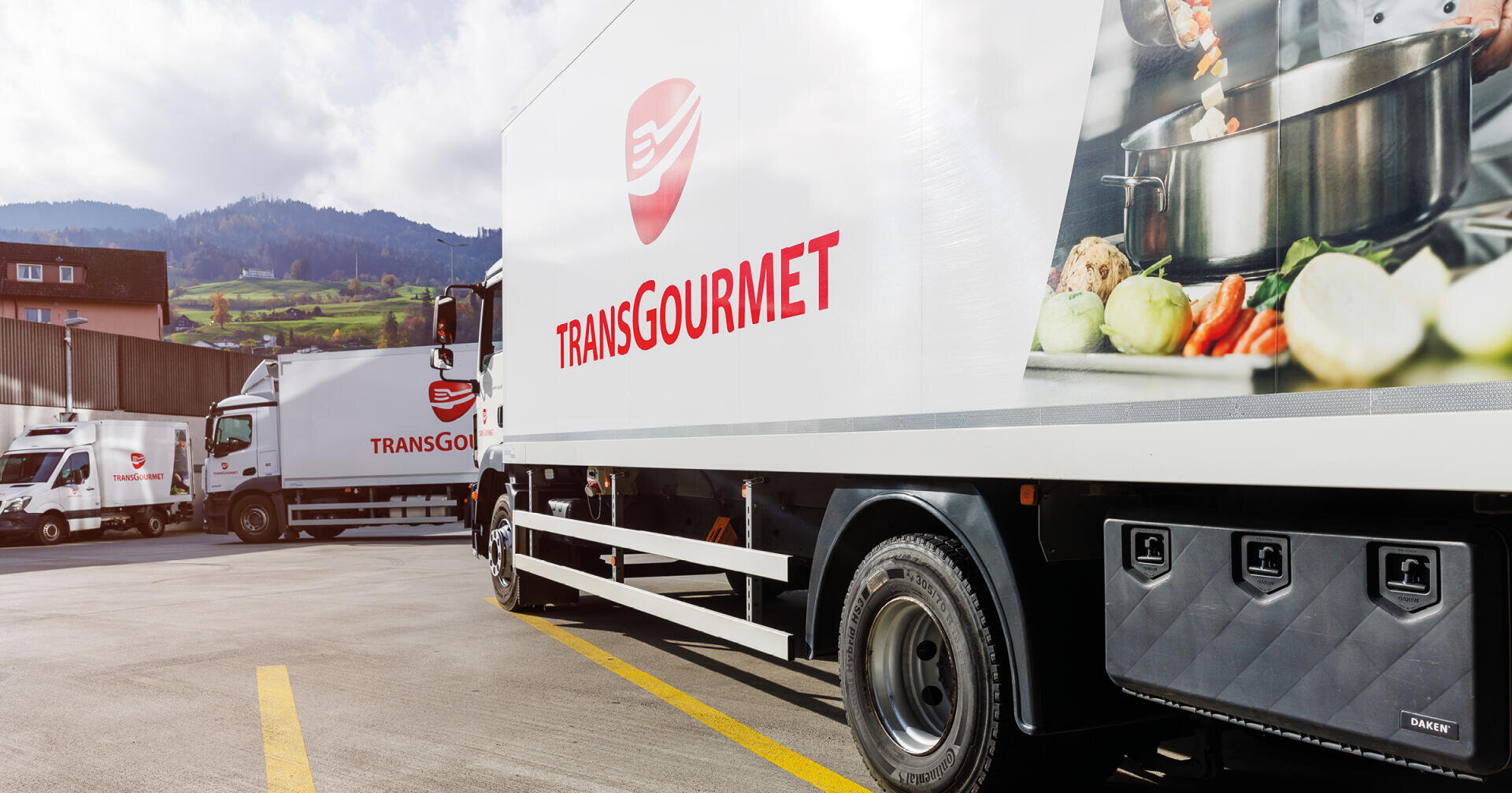 Zwei Lieferwagen von Transgourmet standen auf einem Parkplatz; auf ihren Seitenwänden waren das Firmenlogo und eine Grafik zur Zubereitung von Speisen zu sehen.