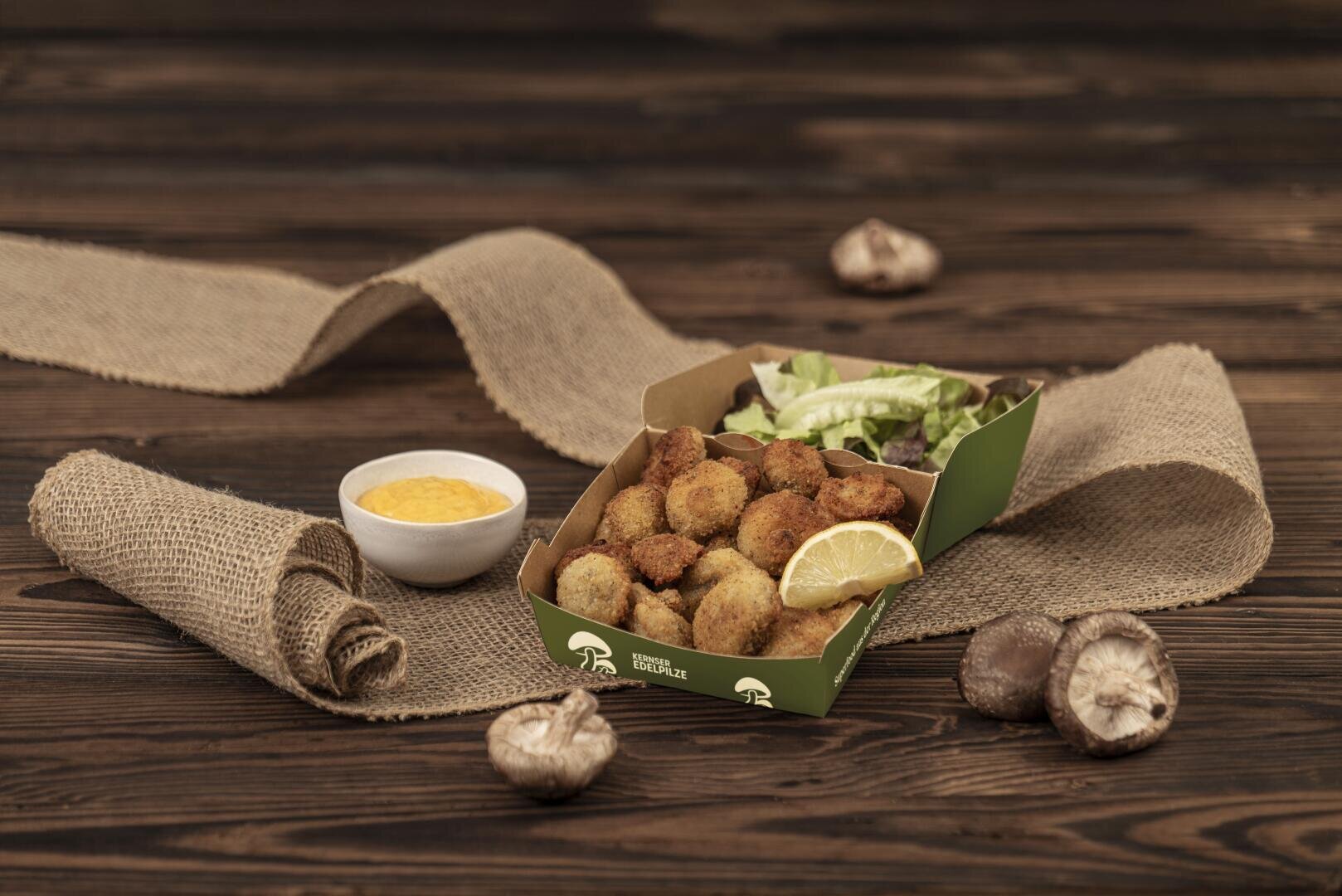 Eine Schale mit panierten, frittierten Pilzen, dazu eine Zitronenscheibe, ein Blattsalat und eine Schüssel mit gelber Dip-Sauce auf einem rustikalen Holztisch, der mit Sackleinen bedeckt ist und auf dem ganze Pilze liegen.