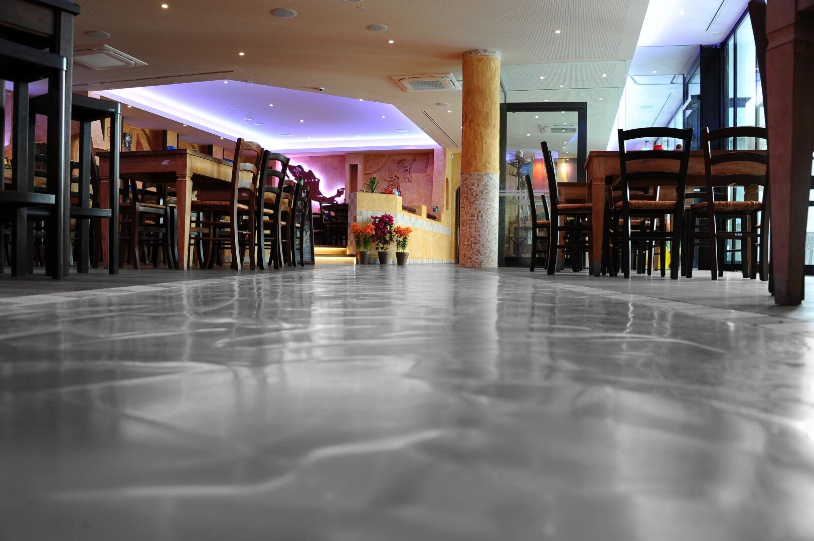 Ein modernes Restaurantinterieur mit Holztischen und -stühlen, einem glänzenden, spiegelnden Boden, violetten Deckenleuchten und einer Vase mit Blumen in der Mitte des Raums.
