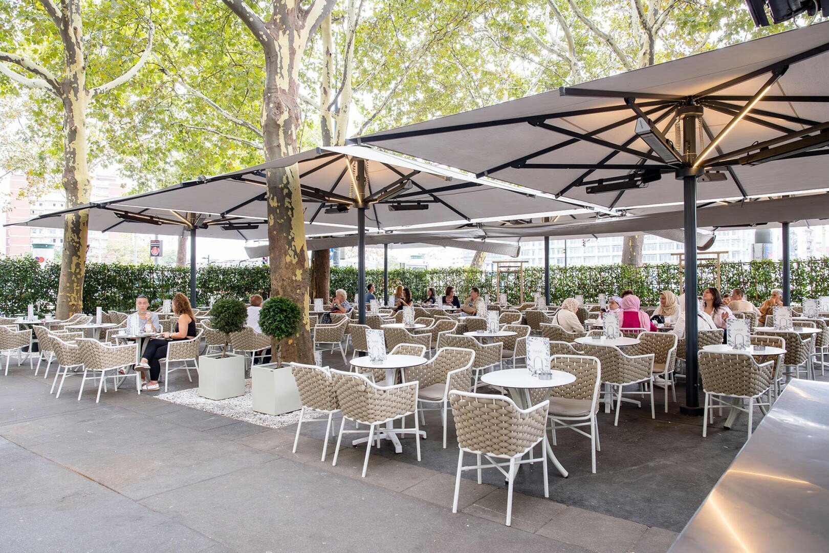 Terrasse eines Restaurants im Freien mit weißen Tischen und Stühlen, großen Sonnenschirmen, die Schatten spenden, und Laubbäumen über den Köpfen. Mehrere Menschen sitzen dort und essen, umgeben von Grün und weitem Freiraum.