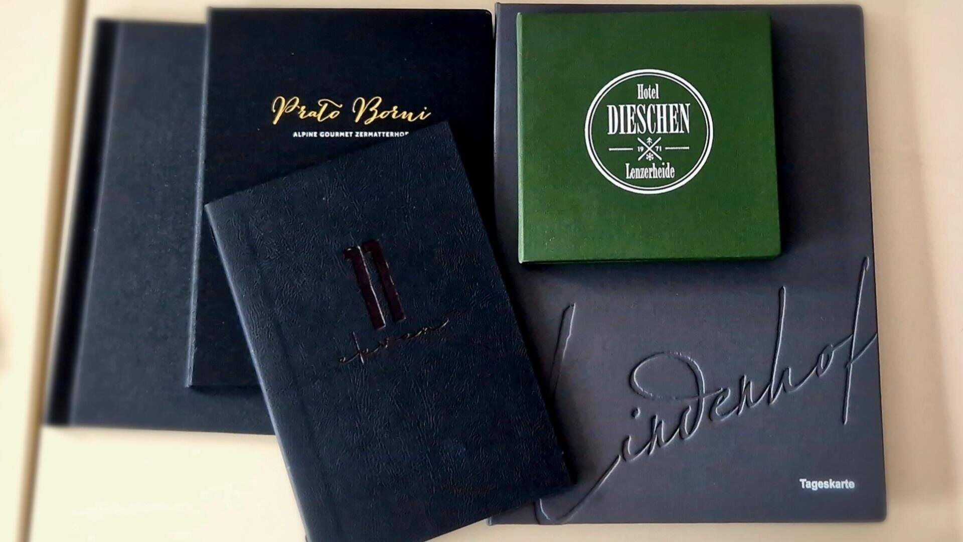 Vier Speisekarten sind übereinander gestapelt und überlappen sich. Die Einbände sind schwarz oder grün, und die Namen „Proto Beni“, „DIESCHEN“ und „Löwenhof“ sind in unterschiedlichen Schriftarten und Stilen zu erkennen.