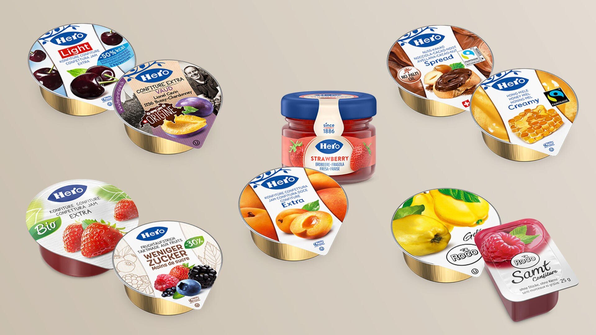 Verschiedene Gläser mit Fruchtkonfitüre und Aufstrichen der Marke „Hero“, darunter Sorten wie Erdbeere, Himbeere, Aprikose, Quitte und weitere, angeordnet vor einem beigen Hintergrund. In der Mitte steht ein Glas Erdbeerkonfitüre.