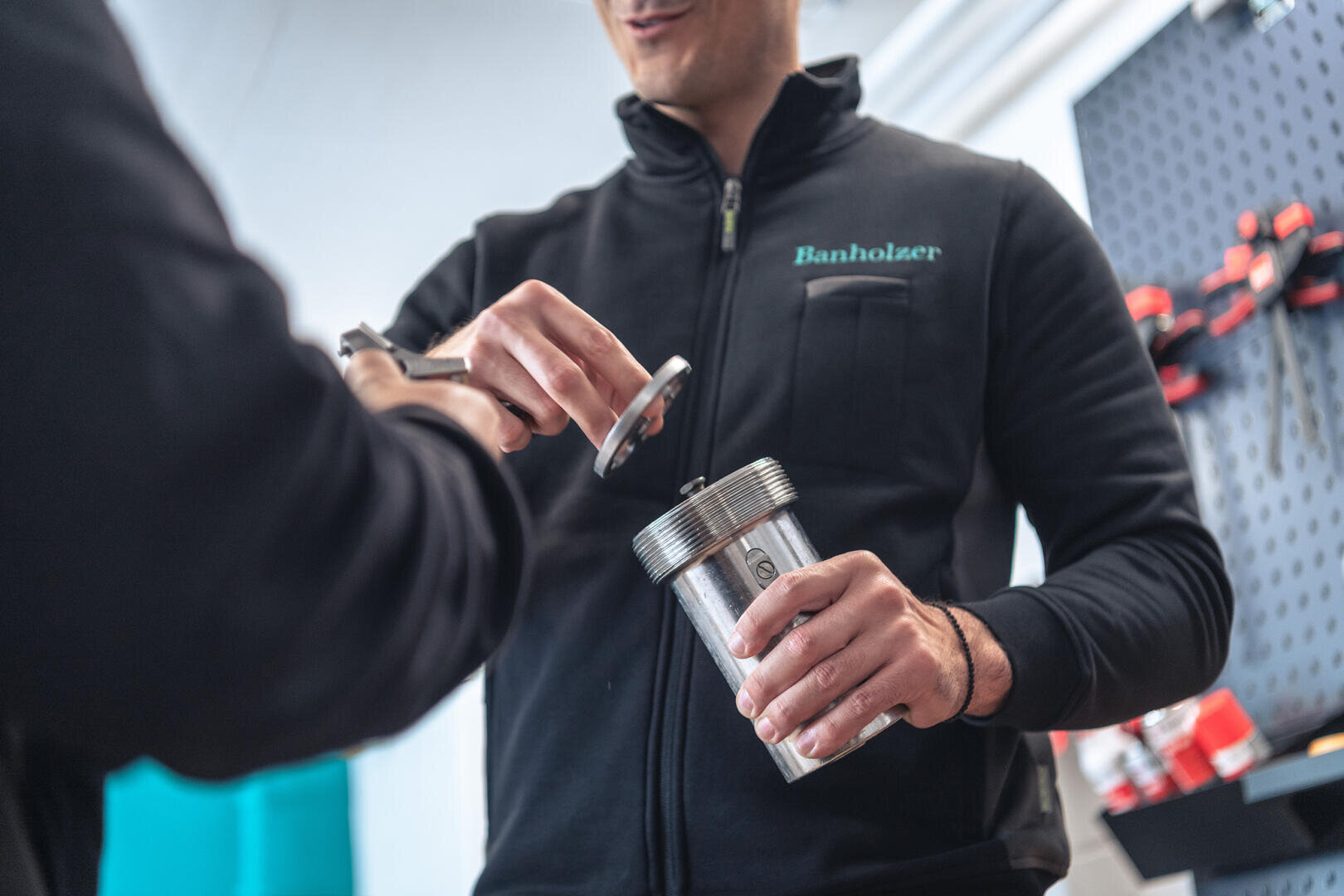 Zwei Personen arbeiten gemeinsam mit Metallwerkzeugen; eine hält einen Metallzylinder fest, während die andere ein rundes Metallteil justiert. Beide sind dunkel gekleidet; eine trägt eine Jacke mit der Aufschrift „Banholzer“. Im Hintergrund sind Regale mit Werkzeugen zu sehen.