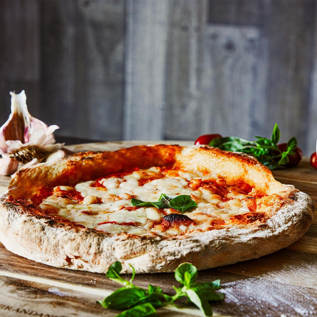Eine Margherita-Pizza mit geschmolzenem Käse und Basilikumblättern auf einem Holzbrett, umgeben von frischem Basilikum, Knoblauch und Tomaten.