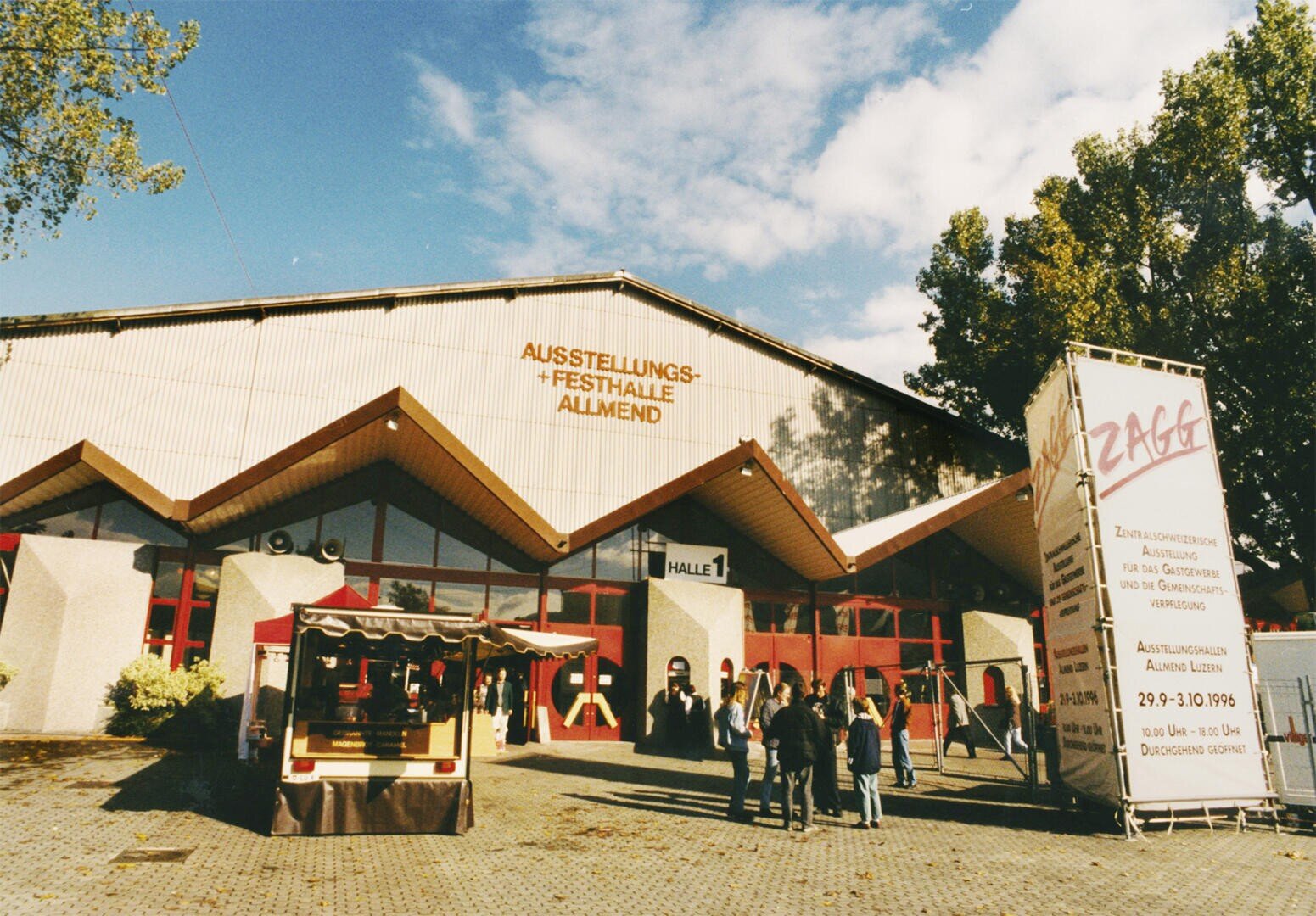Vor einer großen Ausstellungshalle mit der Aufschrift „AUSSTELLUNGS- + FESTHALLE ALLMEND“ stehen Menschen. Im Vordergrund sind ein Imbissstand und ein großes Schild mit Informationen zur Veranstaltung zu sehen. Der Himmel ist teilweise bewölkt.