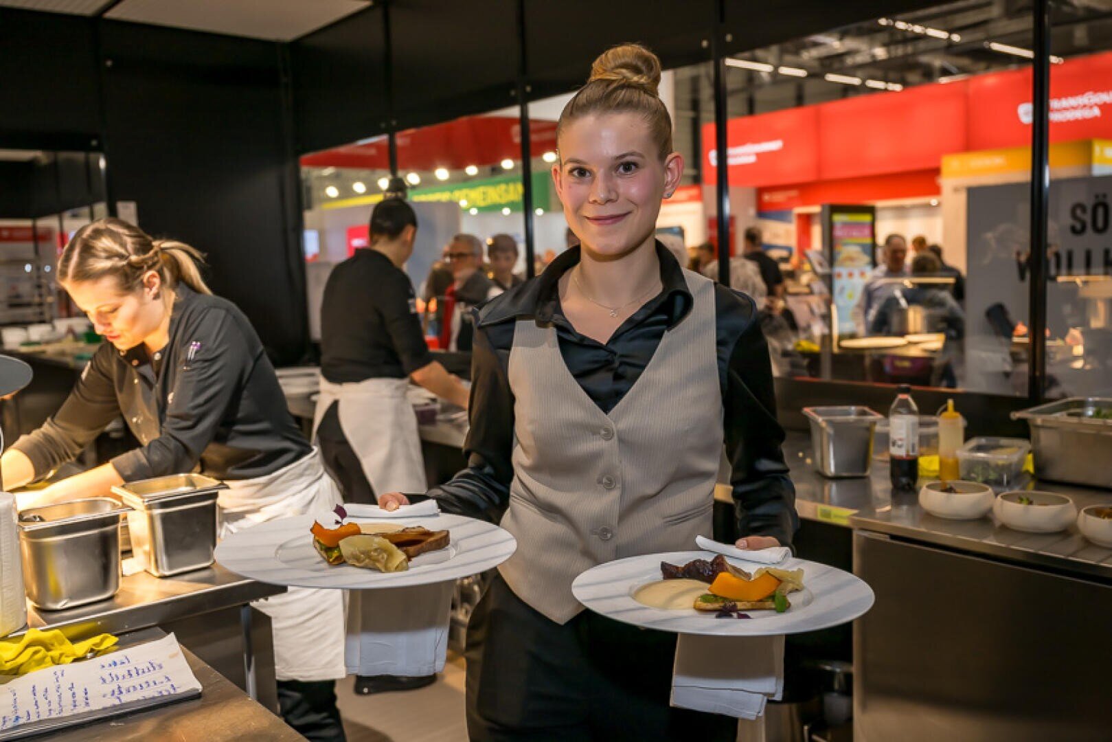 Eine Kellnerin in einer beigen Weste hält in einer geschäftigen, modernen Restaurantküche zwei Teller mit Gourmetgerichten in der Hand. Im Hintergrund bereiten Köche unter grellem Licht Gerichte zu.