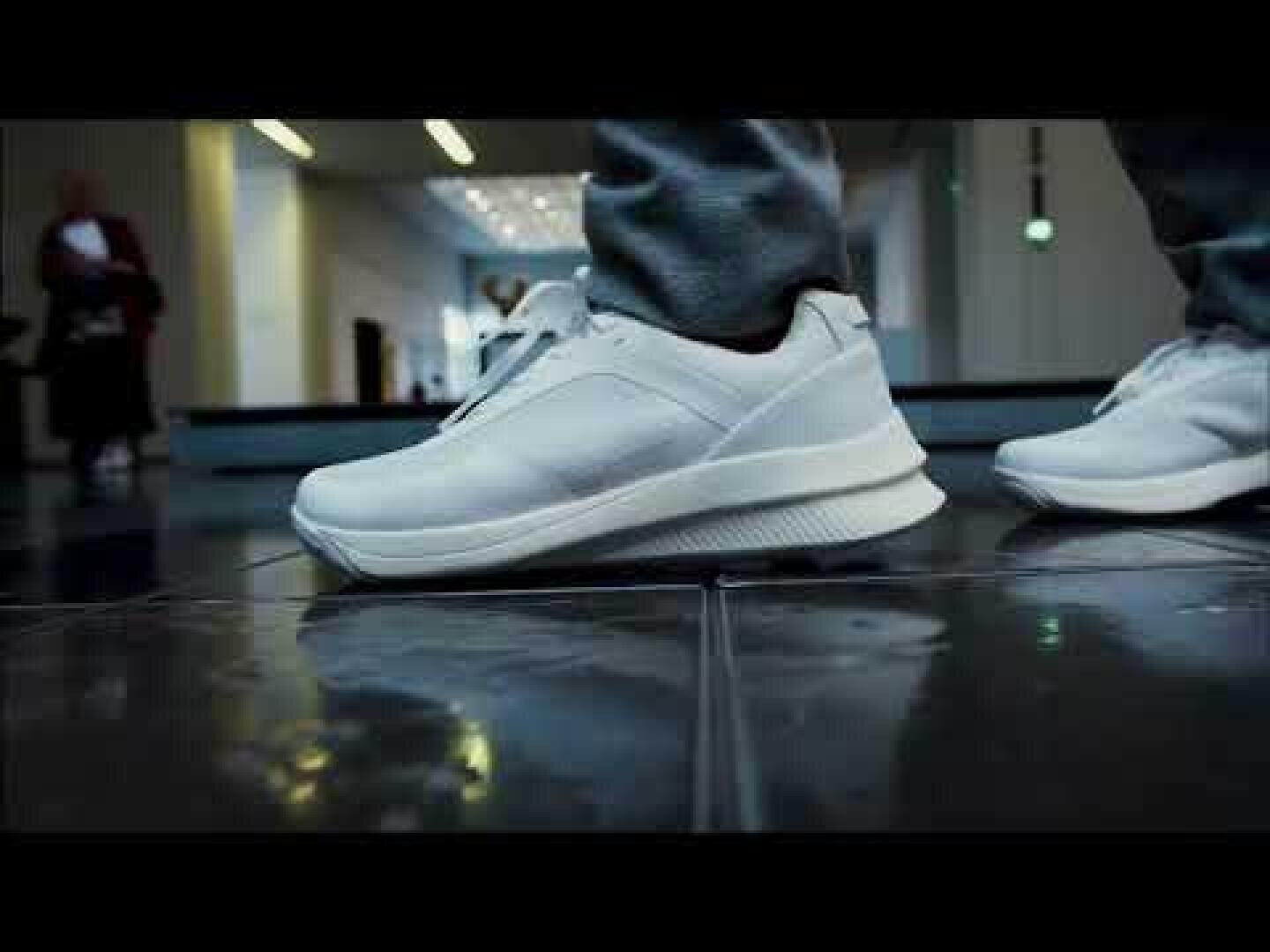 Nahaufnahme einer Person, die weiße Turnschuhe und eine graue Hose trägt und in einem Innenraum auf einem glänzenden Fliesenboden steht, im Hintergrund sind verschwommene Gestalten und Lichter zu sehen.