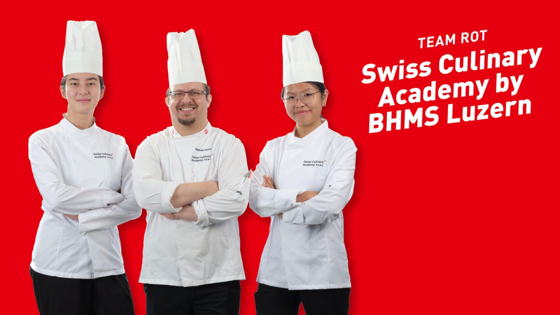 Drei Köche in weissen Uniformen und hohen Kochmützen stehen mit verschränkten Armen vor einem Schriftzug mit der Aufschrift Team Rot, Swiss Culinary Academy by BHMS Luzern.