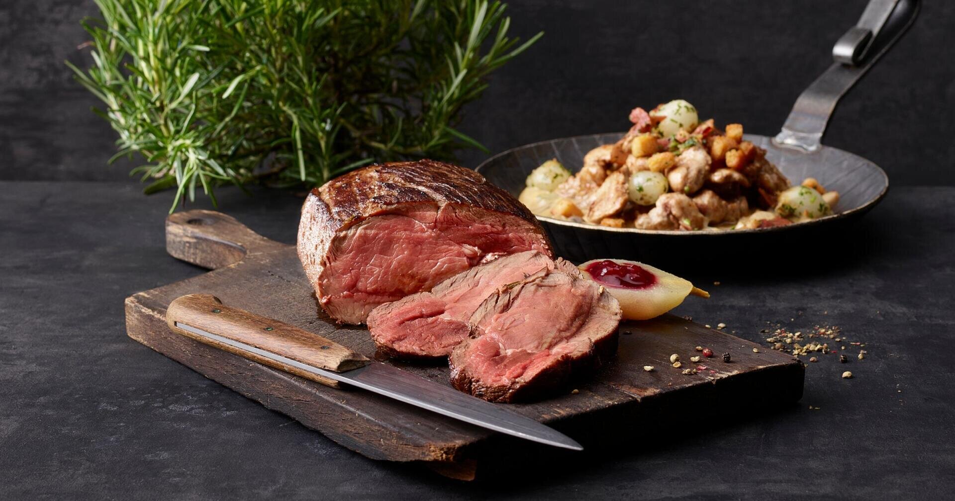 Ein in Scheiben geschnittenes Roastbeef auf einem hölzernen Schneidebrett mit einem Messer, garniert mit Kräutern und Soße; im Hintergrund steht eine Pfanne mit Bratkartoffeln und Croutons.