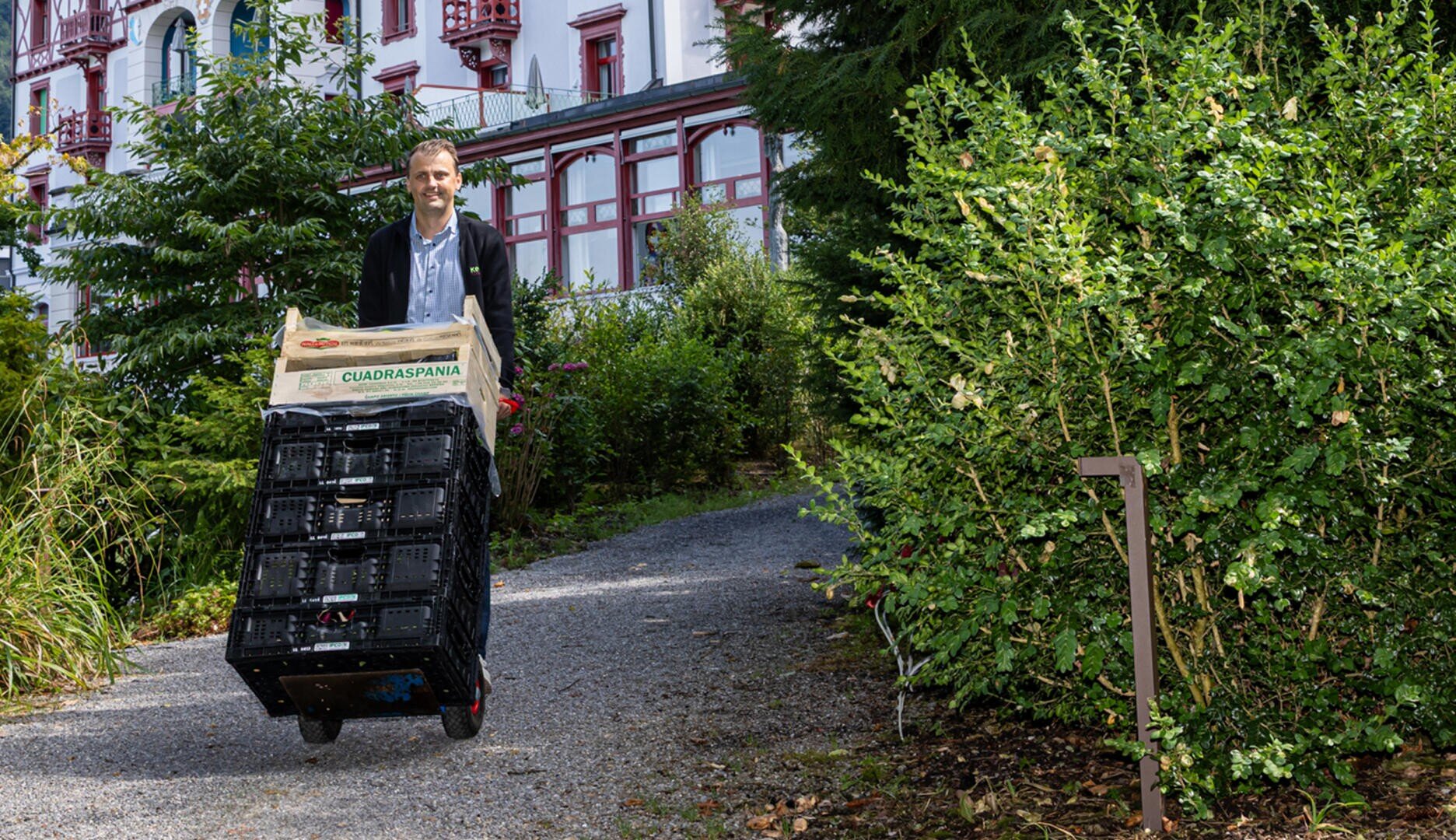 Ein Mann geht auf einem Schotterweg durch einen Garten und schiebt dabei einen Karren, auf dem schwarze Kisten und eine Schachtel mit der Aufschrift „Cuadraspania“ gestapelt sind. Im Hintergrund ist ein großes Gebäude mit rot-weißen Zierleisten zu sehen.