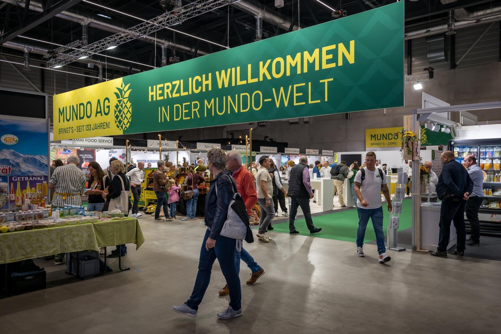Menschen, die durch eine hell erleuchtete Messehalle unter einem großen grünen Banner mit der Aufschrift Herzlich willkommen in der Mundo-Welt gehen.