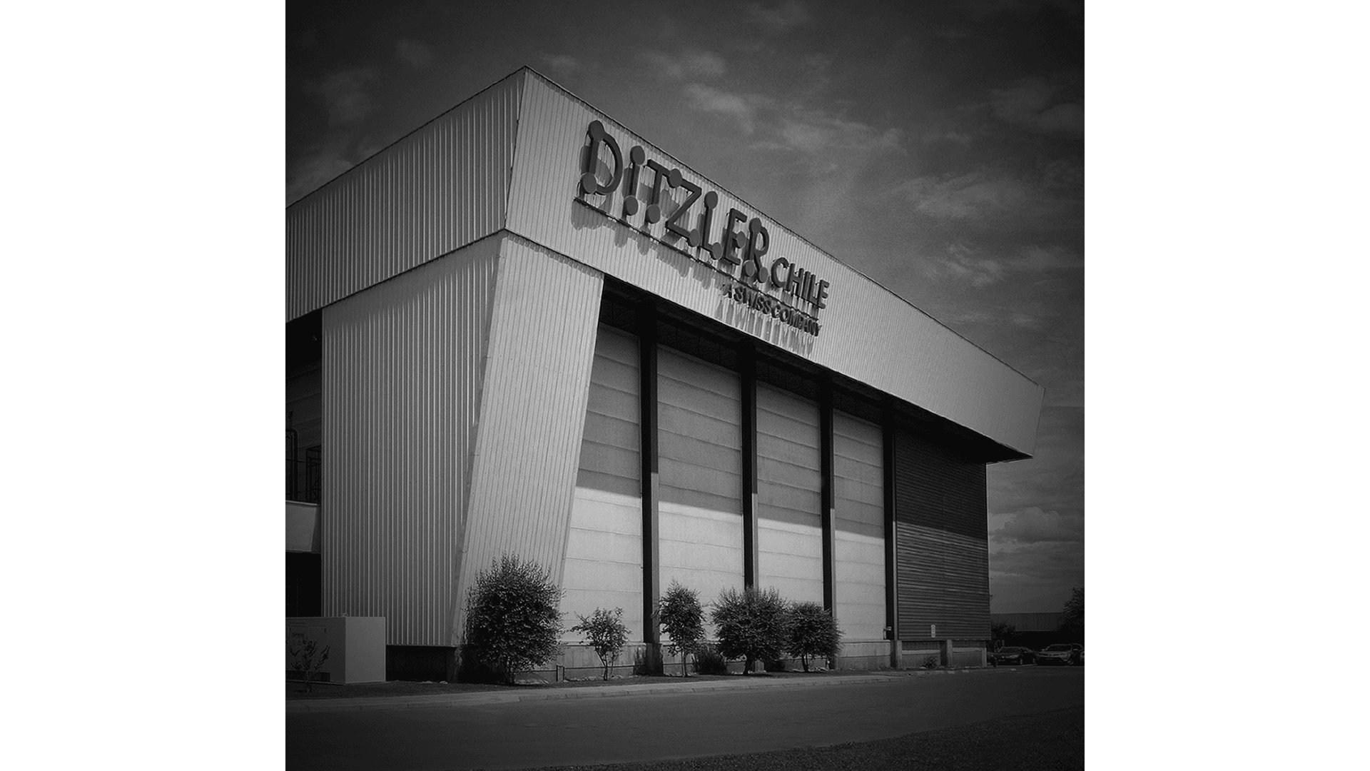 Ein großes Gebäude im Industriestil mit hohen, weißen Metallplatten und schrägen Wänden. Auf der Fassade steht DIZZLER CABLE MANUFACTURING. Mehrere kleine Bäume und Sträucher säumen die Vorderseite des Gebäudes unter einem teilweise bewölkten Himmel.