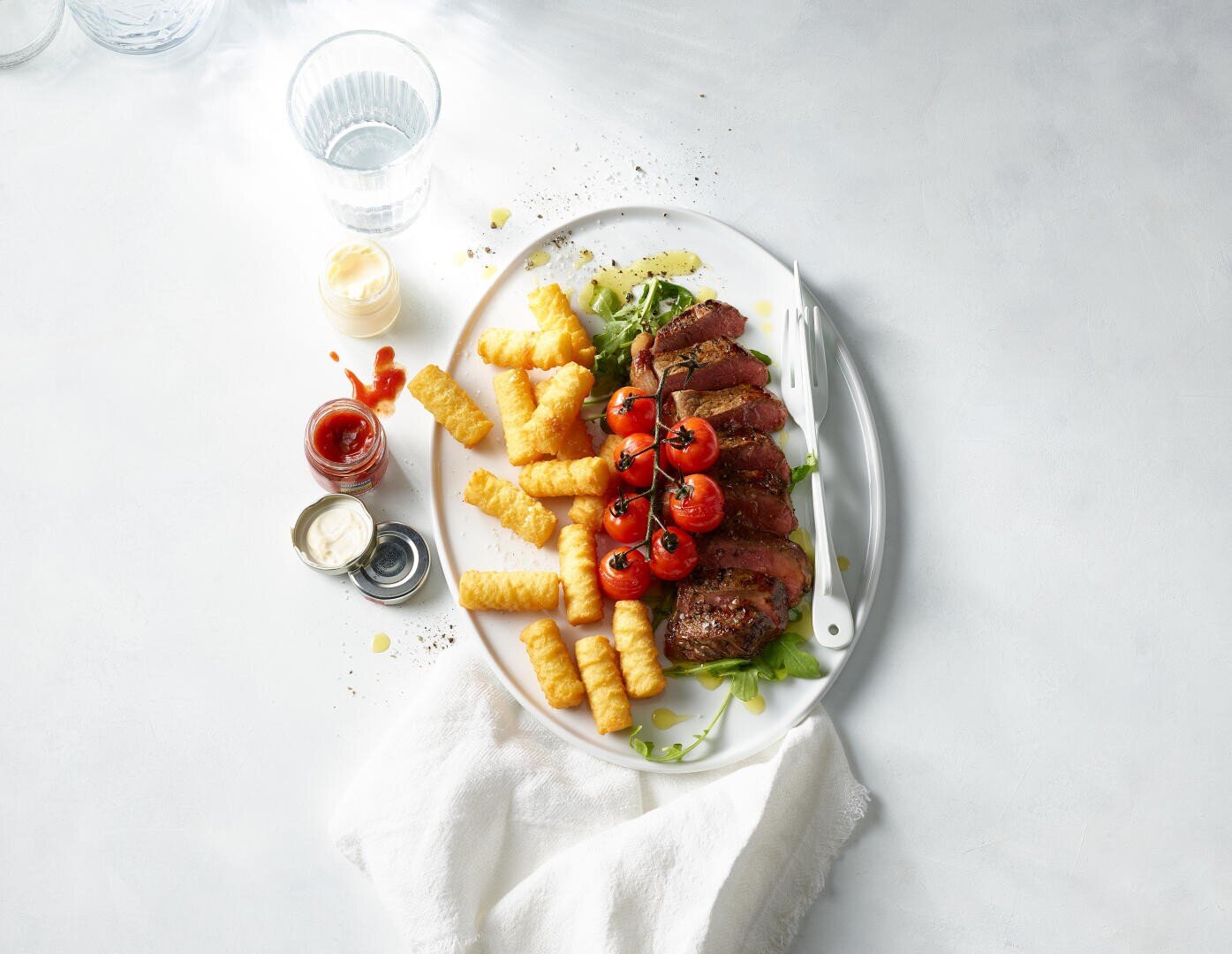 Ein weißer Teller mit Steakscheiben, Kirschtomaten am Stiel, gewellten Pommes frites und Blattgemüse. Neben dem Teller stehen drei kleine Soßenschälchen und eine Gabel. Ein Glas Wasser und eine weiße Serviette liegen daneben.