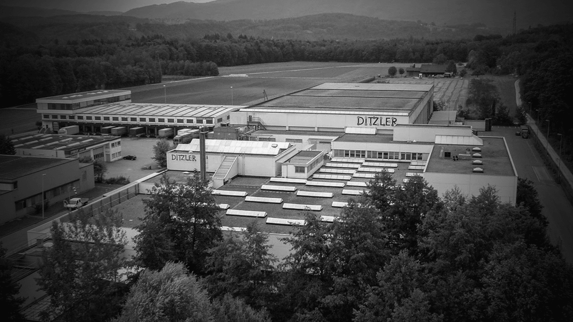 Luftaufnahme eines Industriekomplexes, umgeben von Bäumen und Feldern, mit mehreren großen Gebäuden mit der Aufschrift DITZLER und Hügeln im Hintergrund. Das Bild ist in schwarz-weiß gehalten.