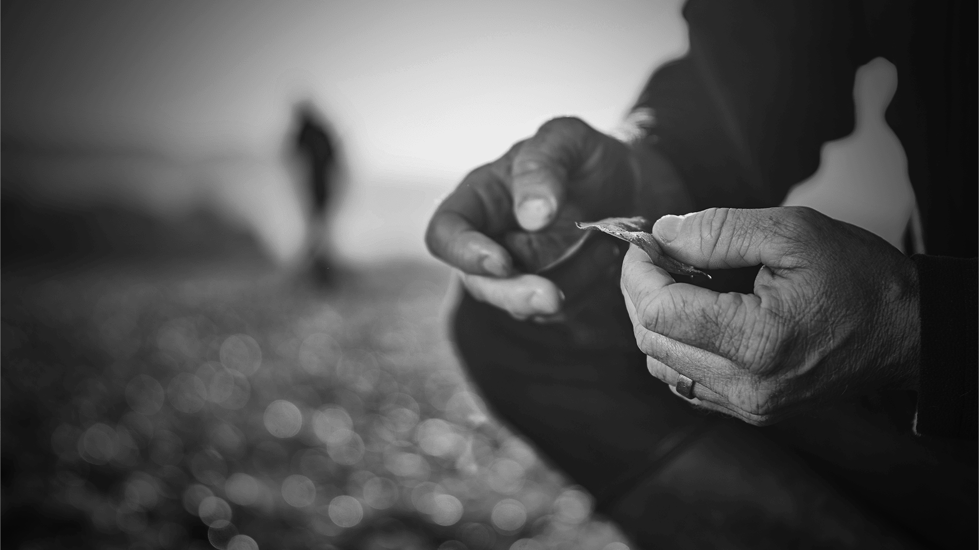 Nahaufnahme einer Person, die im Freien einen kleinen Gegenstand in der Hand hält, mit einer anderen Figur, die im Hintergrund verschwommen ist. Das Bild ist in Schwarz-Weiß gehalten, was eine kontemplative Stimmung erzeugt.
