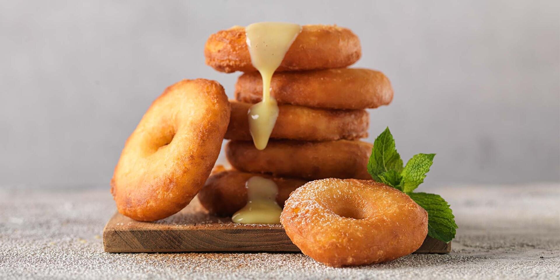 Ein Stapel goldbrauner Donuts auf einem Holzbrett, von denen süße Glasur herabtropft, garniert mit einem Zweig frischer Minze. Ein Donut liegt ganz vorne und ist leicht mit Zucker bestäubt.