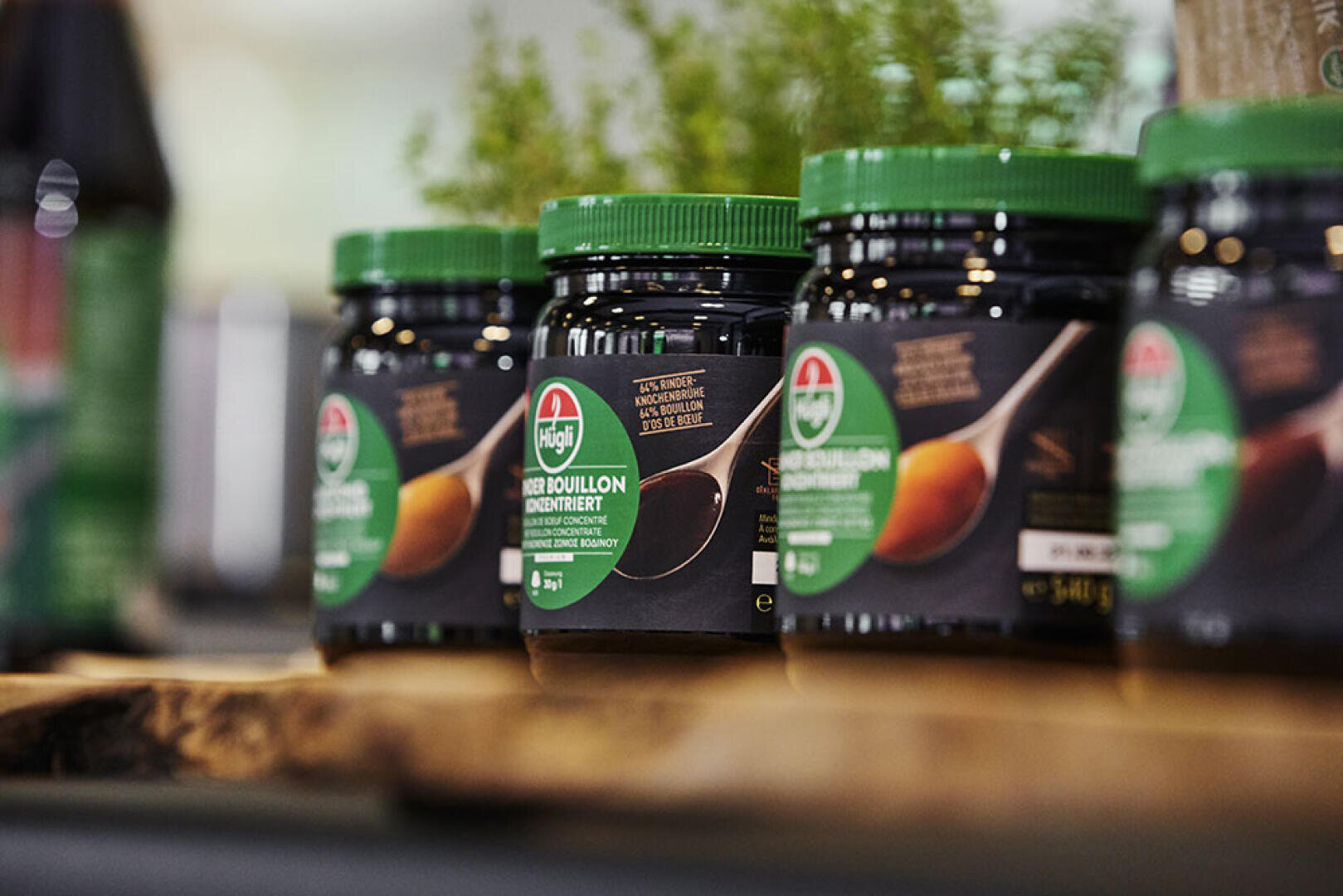Eine Reihe von Gläsern mit grünen Deckeln und grünen Etiketten, die auf einer Holzfläche ausgestellt sind. Auf den Gläsern steht „Bio-Brühe“. Im Hintergrund ist etwas Grün zu sehen.