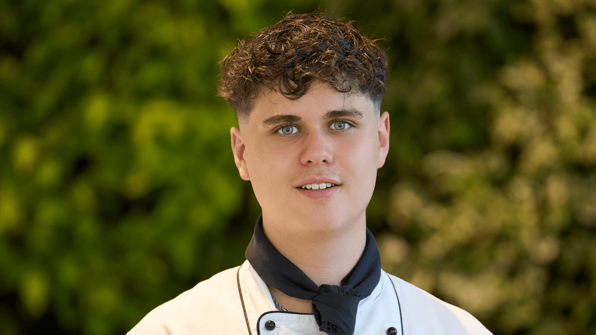 Ein junger Koch in weißer Uniform und mit schwarzem Halstuch blickt in die Kamera.