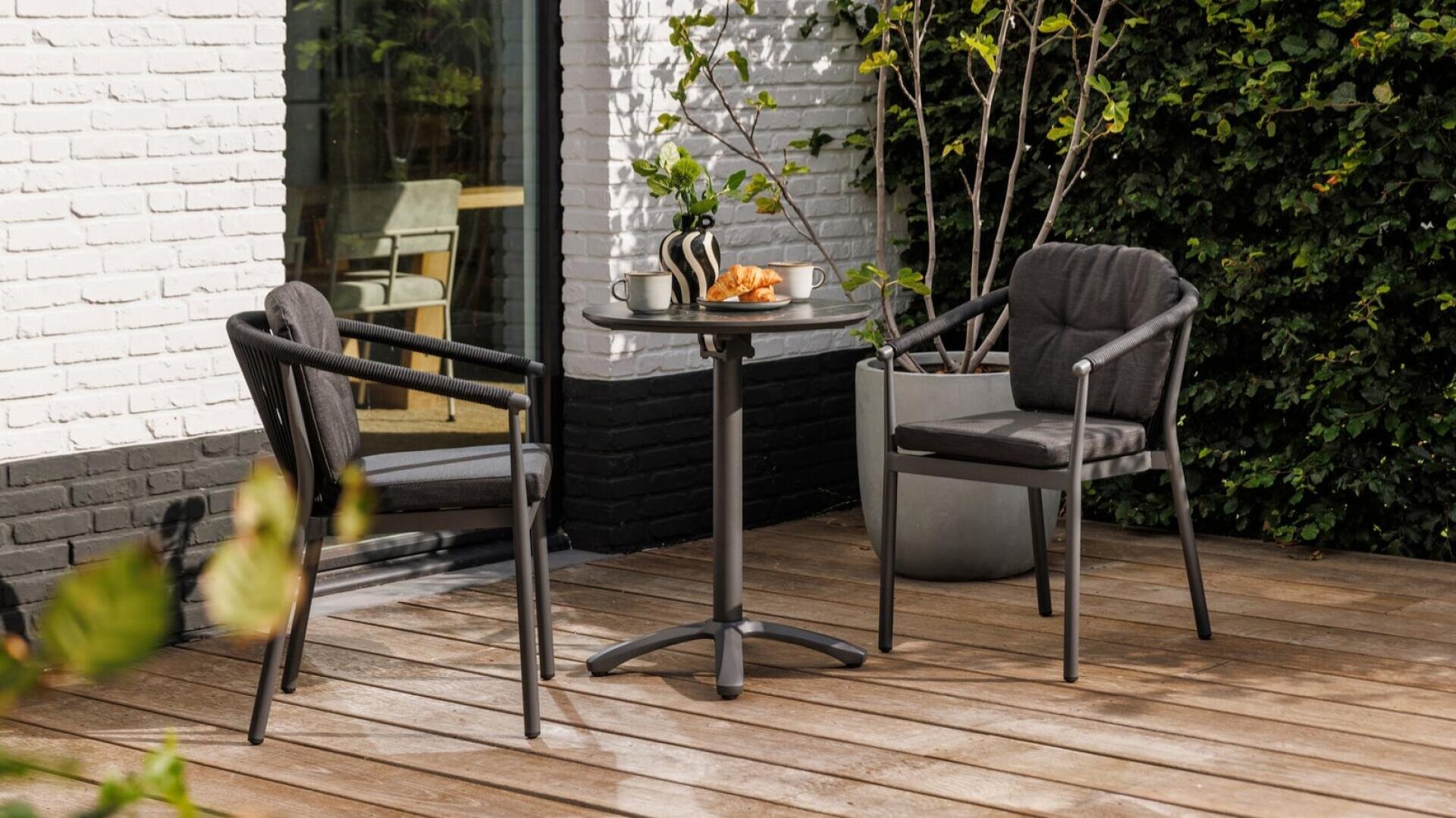 Eine kleine Terrasse mit zwei schwarzen Stühlen mit Polstern und einem runden Tisch, auf dem eine gestreifte Vase, eine Tasse und ein Teller mit Croissants stehen; die Terrasse befindet sich auf einem Holzbelag neben einem weißen Backsteingebäude, in dessen Nähe Pflanzen stehen.