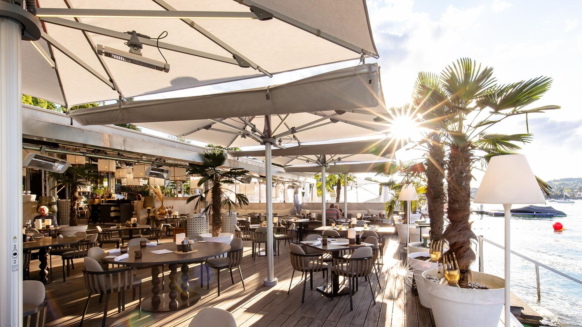 Ein Restaurant im Freien mit Palmen, modernen Tischen und Stühlen sowie großen Sonnenschirmen. Das Sonnenlicht scheint durch das Blätterdach, von wo aus man auf das Ufer mit Booten und eine malerische Aussicht blickt.