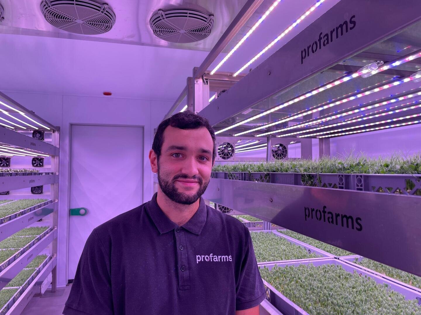 Ein Mann, der ein Profarms-Poloshirt trägt, steht in einer vertikalen Indoor-Farm, in der Reihen von Microgreens unter violetten LED-Wachstumslampen stehen.