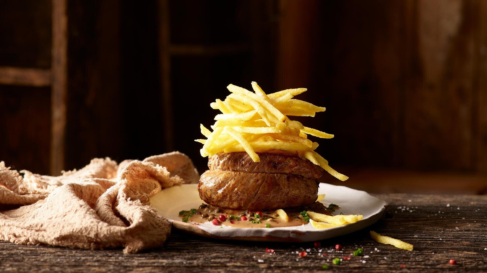Auf einem rustikalen Holztisch liegt ein Steak auf einem weißen Teller, darauf ein großzügiger Haufen dünner, knuspriger Pommes frites, umgeben von Kräutern und Gewürzen.